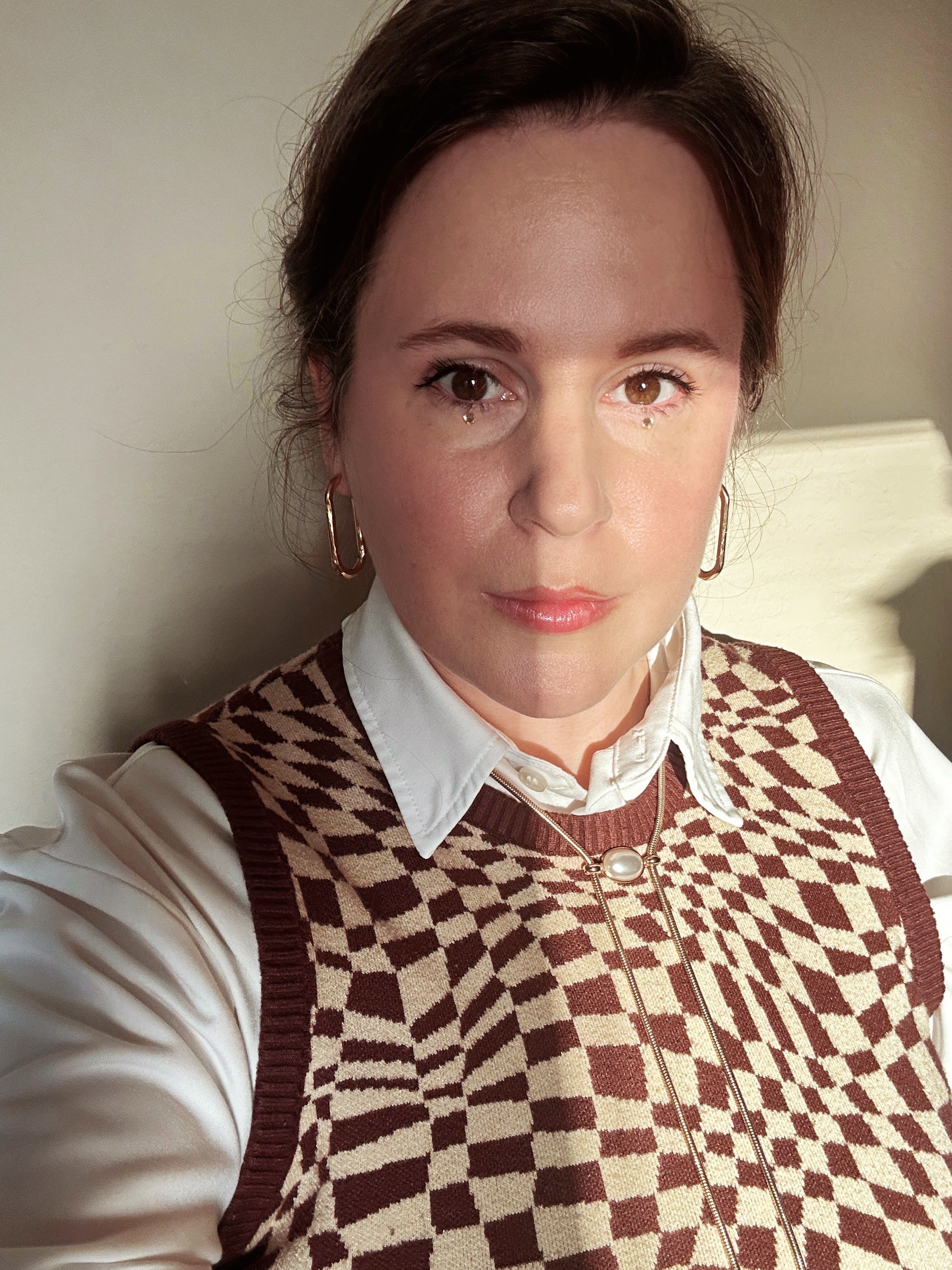 Person wearing a patterned sweater vest over a white shirt, with large hoop earrings and a necklace with a round pendant.
