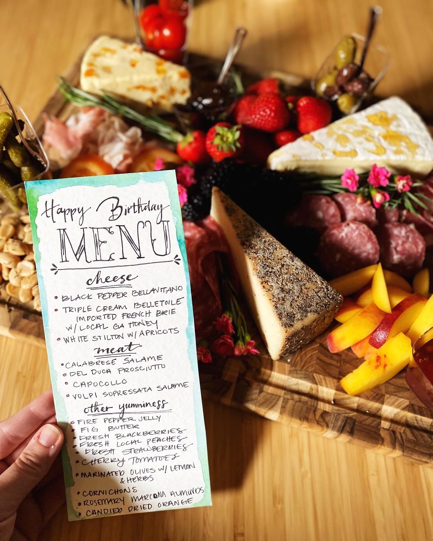 Close-up of a charcuterie board with various cheeses, meats, fruits, and pickles, accompanied by a handwritten menu. The menu lists items such as Black Pepper Bellavitano, Triple Cream Belletoile, Calabrese Salame, and other delicacies. The board is decorated with fresh ingredients like strawberries, blackberries, and herbs.