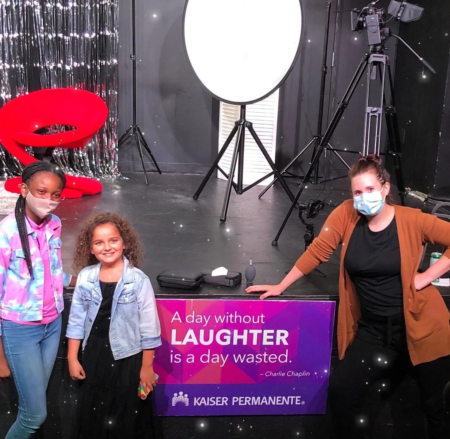 Two young girls and a woman, all smiling, pose in front of a purple sign with a Charlie Chaplin quote about laughter. The woman and one girl wear masks. The setting includes a red chair, camera equipment, and a silver backdrop, suggesting a studio or