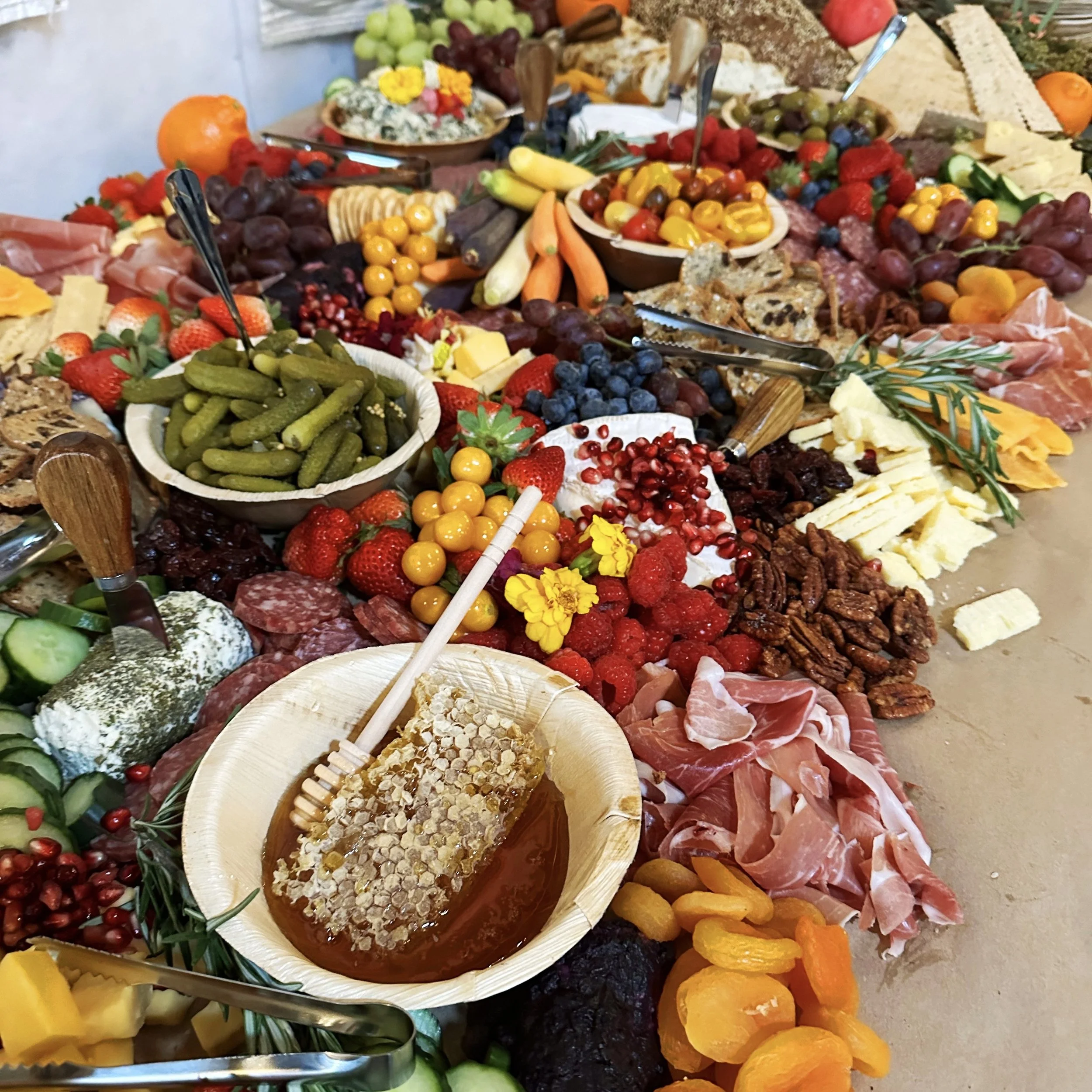 A vibrant charcuterie board featuring a variety of cheeses, cured meats, fruits, nuts, pickles, honeycomb, and crackers. The spread includes strawberries, raspberries, blueberries, grapes, cucumbers, dried apricots, and assorted olives, all artistically arranged.