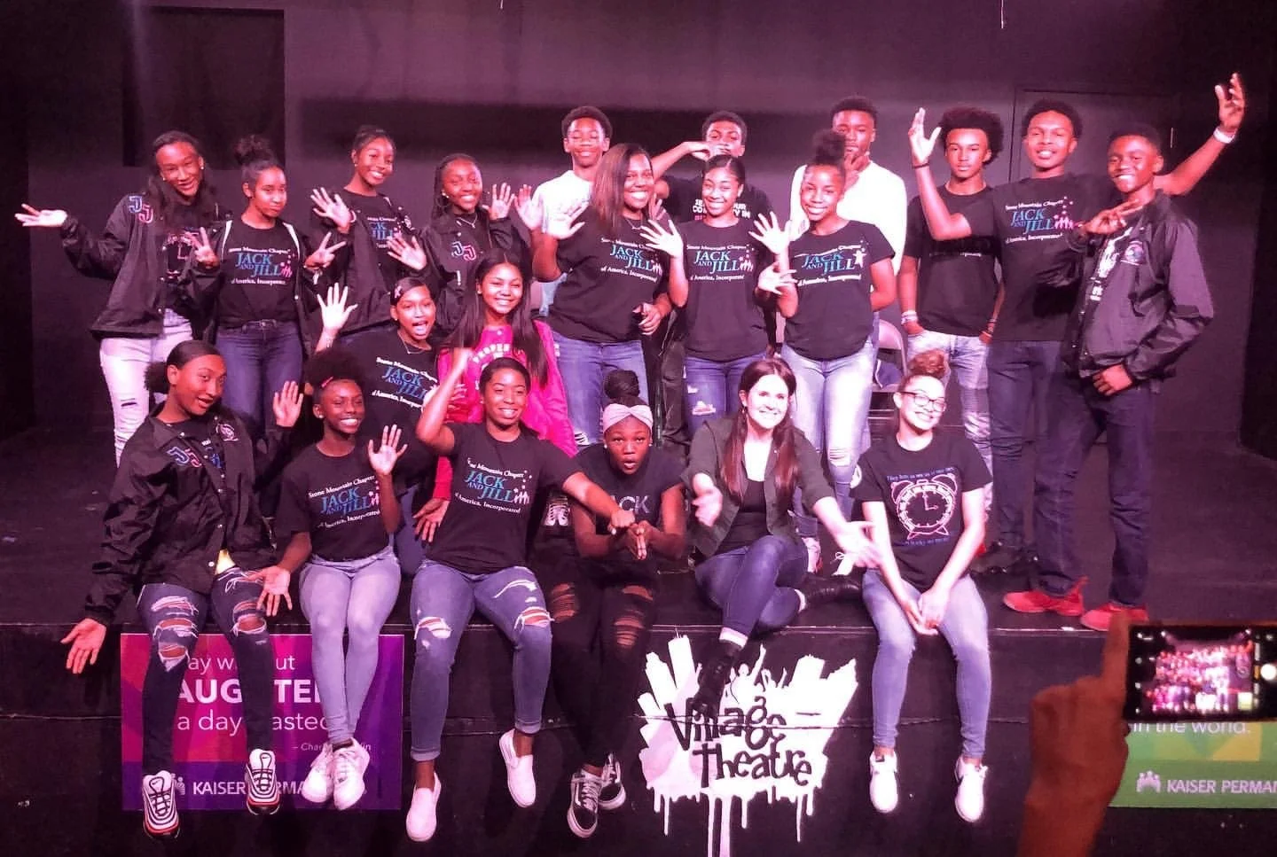 Group of young people posing on stage wearing "Jack and Jill" shirts, with a Village Theatre sign visible.