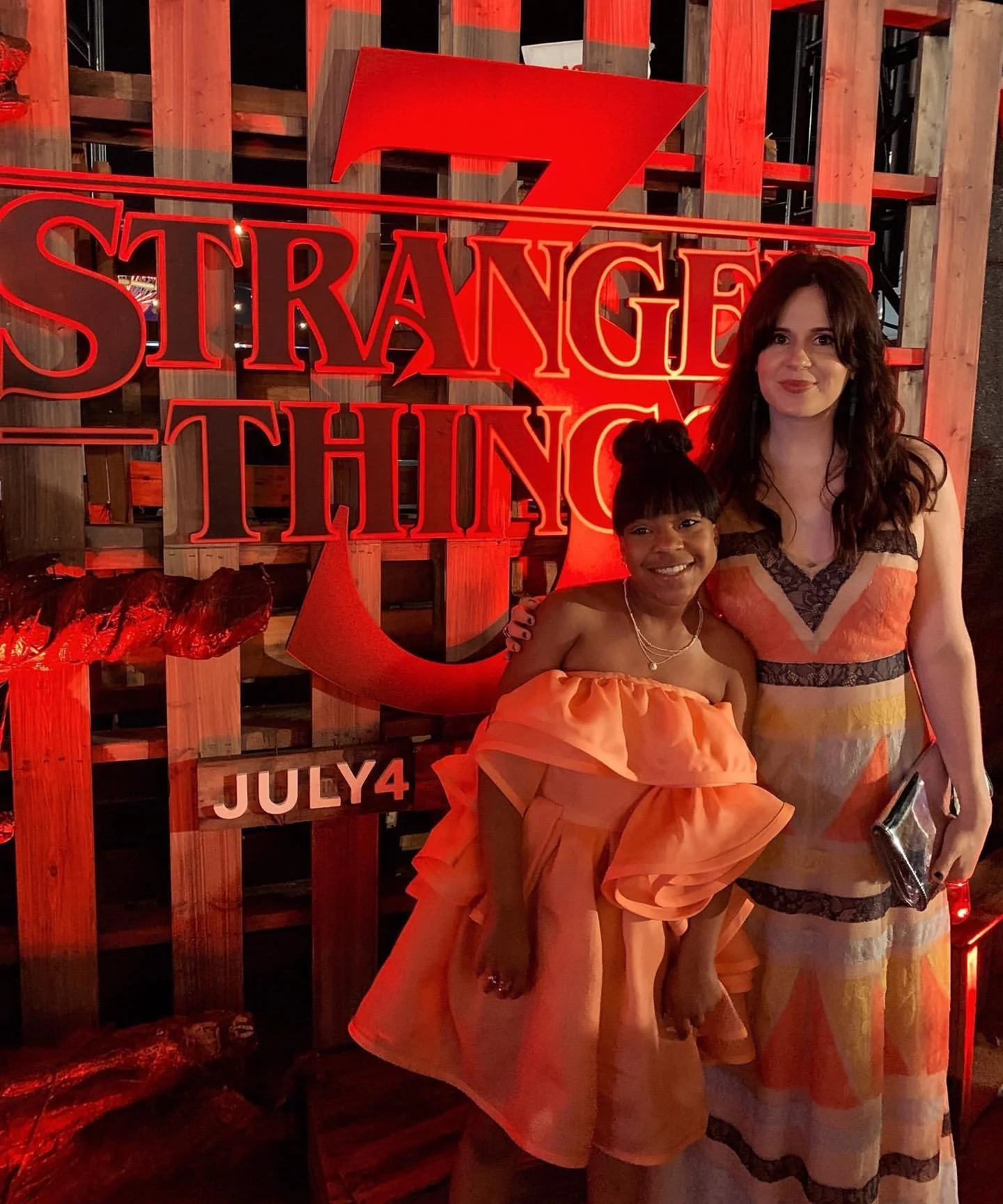 Two people posing in front of a "Stranger Things 3" sign with a red and orange color theme. The sign includes the release date, July 4. Both are wearing stylish dresses suitable for a formal event.