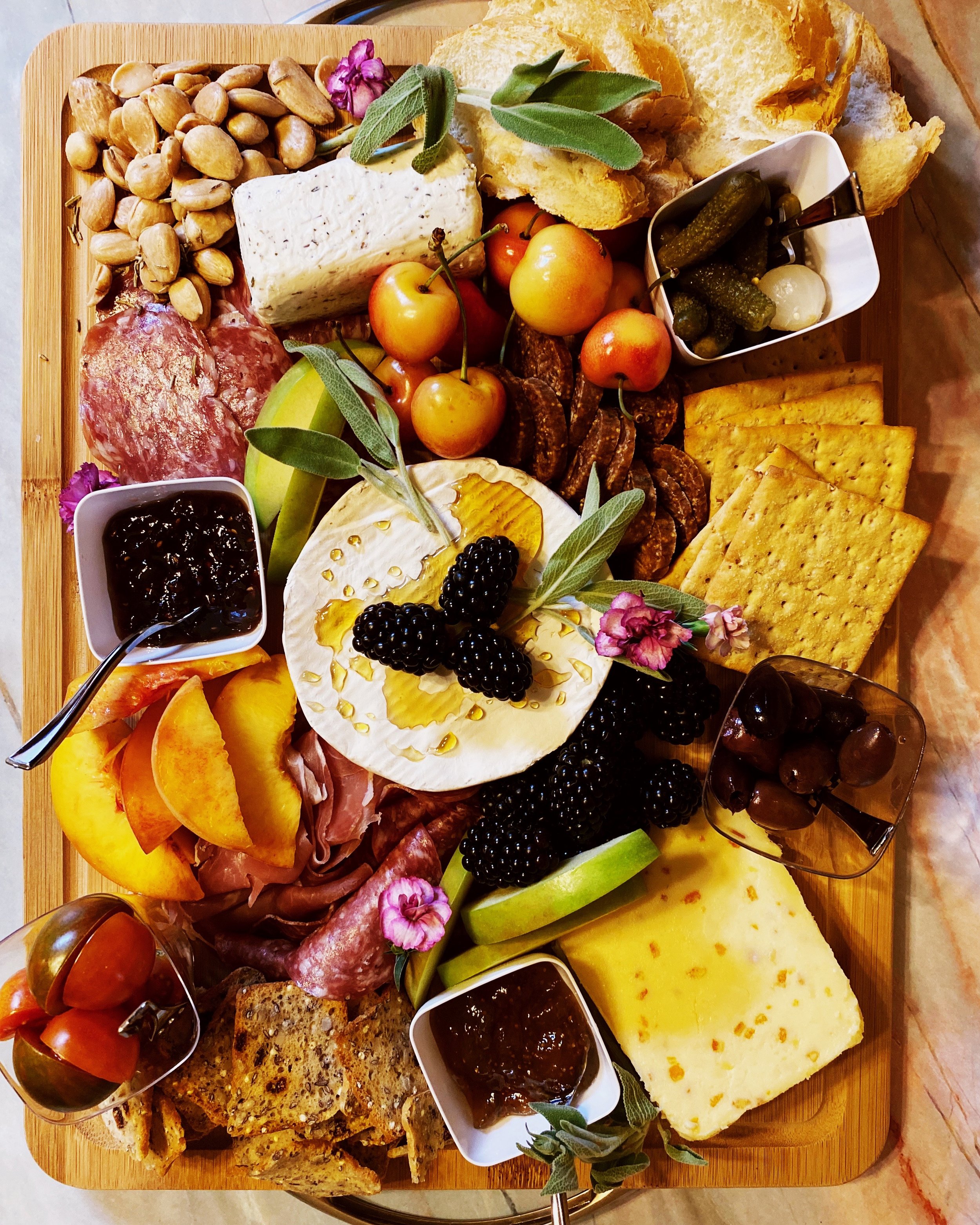 Charcuterie board with cheeses, crackers, almonds, blackberries, cherry tomatoes, peaches, salami, prosciutto, pickles, olives, dried fruit, fig jam, and bread slices, garnished with sage leaves and flowers.