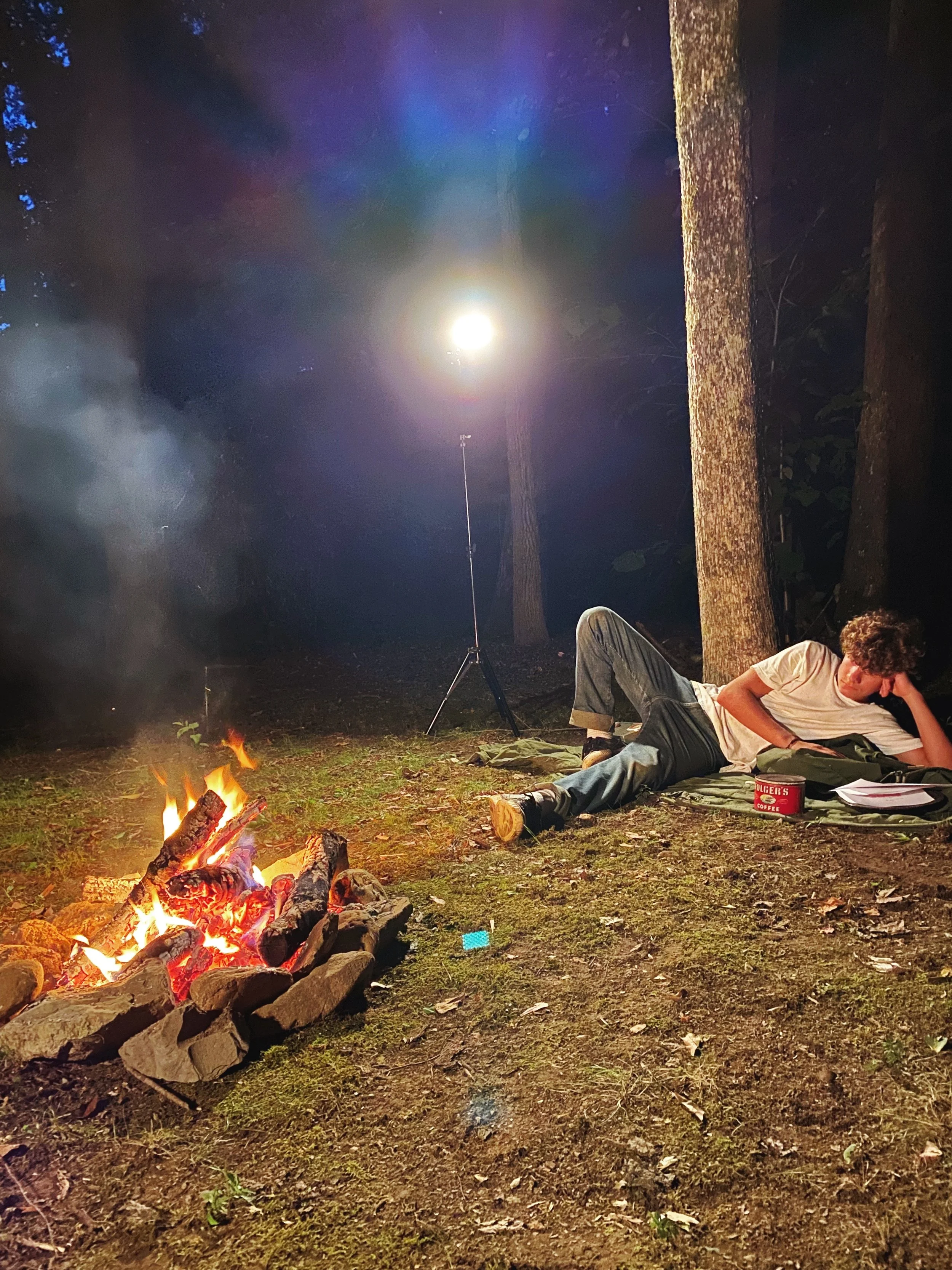 Person lying on a blanket reading near a campfire in a forest with a bright light illuminating the area.