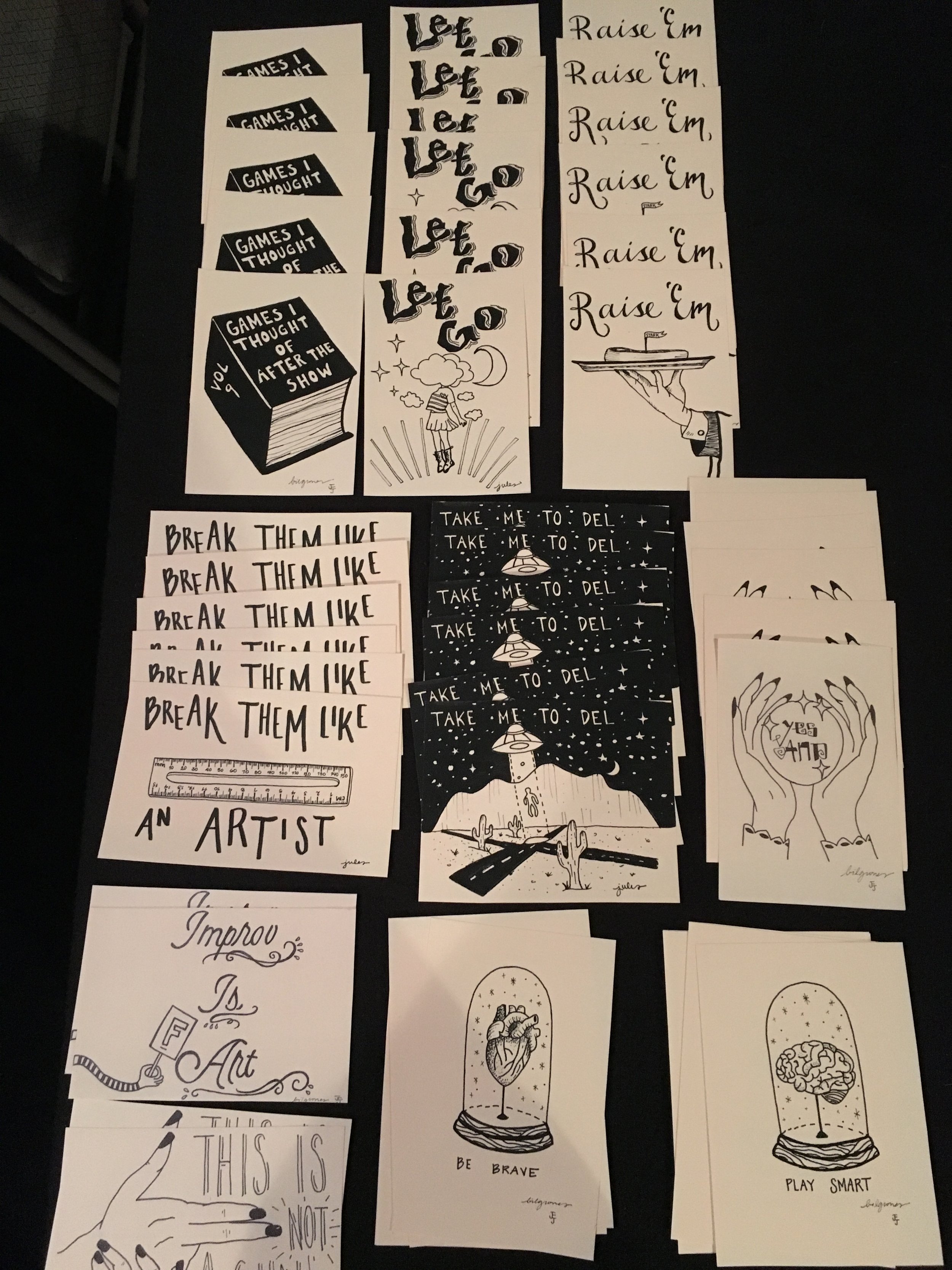 Various black and white illustrations on paper displayed on a table, featuring text like "Let it go," "Raise 'Em," "Break them like an artist," "Improv is art," and "Be brave." Other designs include a book, a hand holding a tray, a girl in the clouds