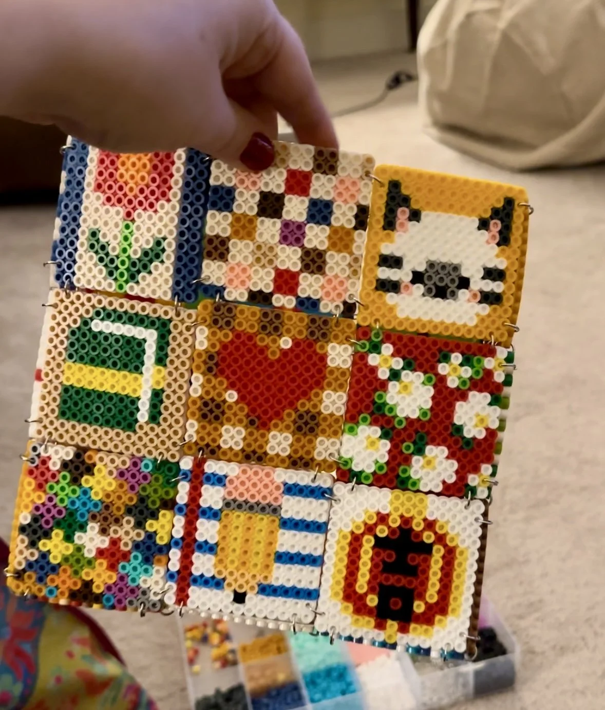 Hand holding a grid of colorful Perler bead designs, including a flower, heart, cat face, and abstract patterns, with a bead storage box in the background.