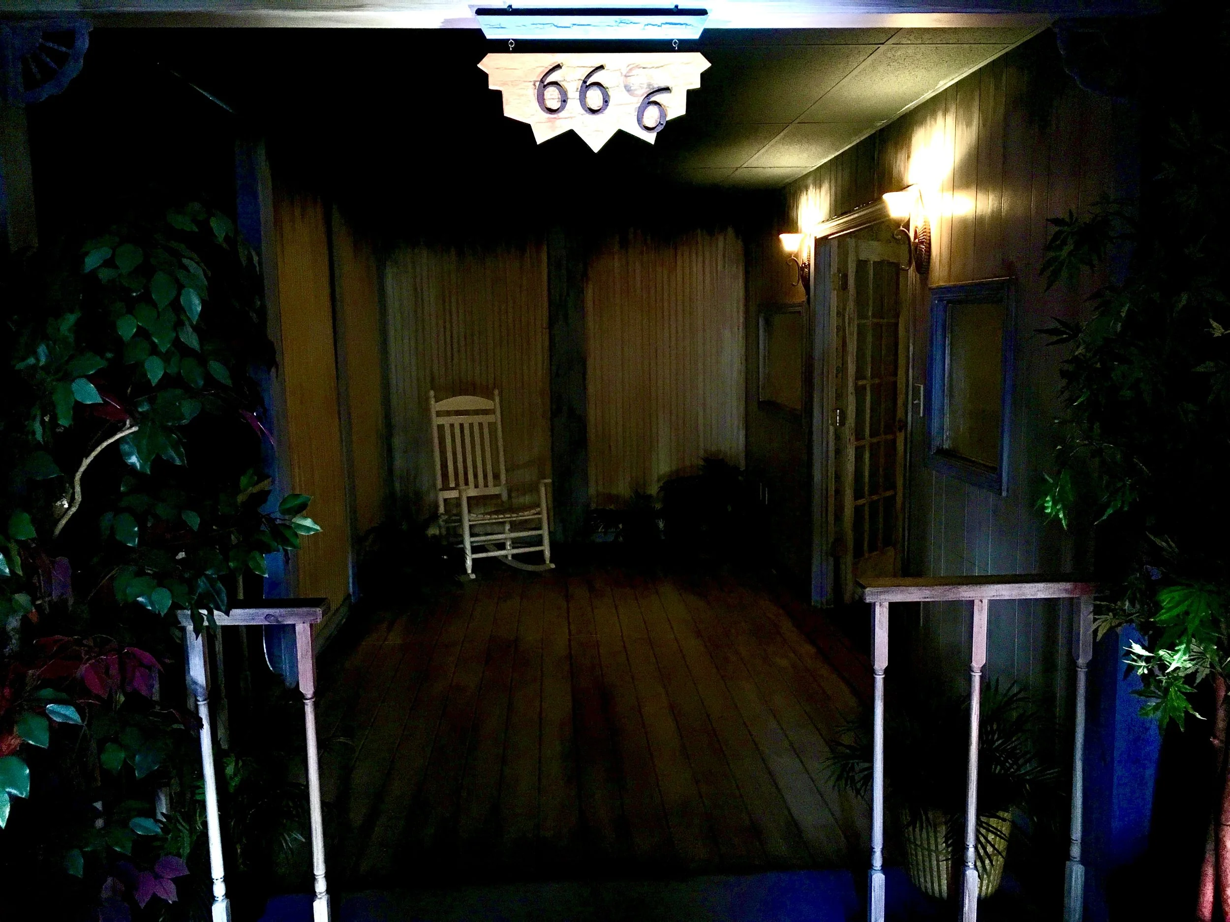 A dimly lit porch with a wooden floor, featuring a white rocking chair and two wall lights. The number '666' is displayed on a wooden sign above the entrance. Potted plants are positioned on either side of the entrance.