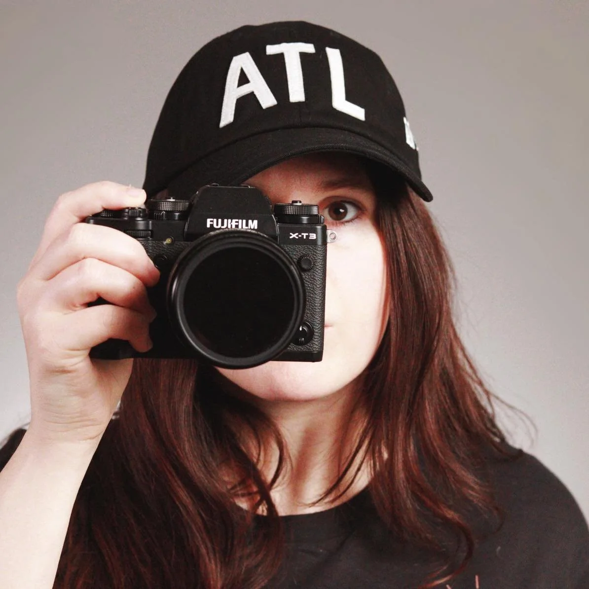 Person wearing a black cap with 'ATL' lettering, holding a Fujifilm X-T3 camera in a self portrait setting.