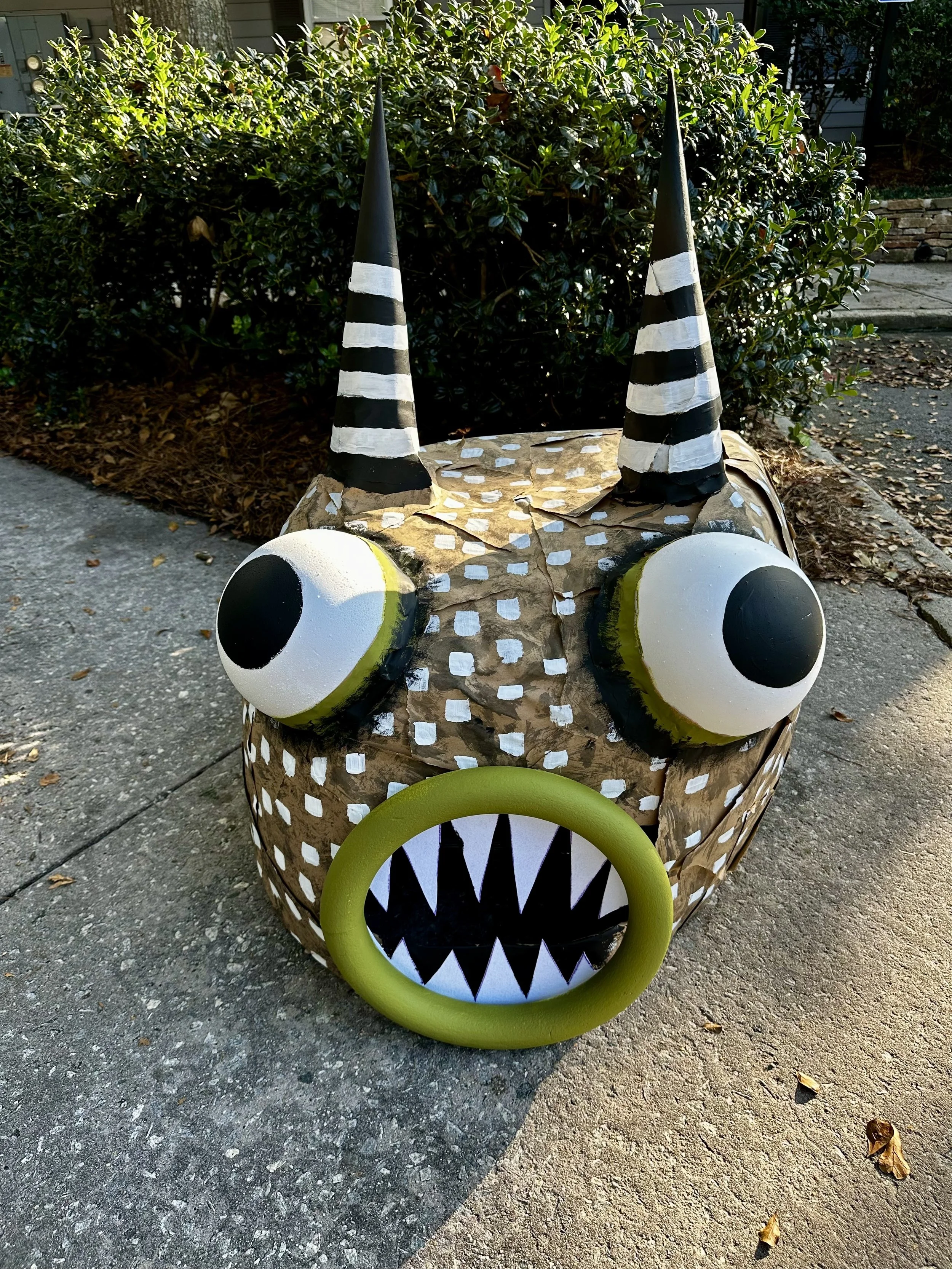 Sculpture of a whimsical monster with large eyes, striped horns, and a circular mouth with pointed teeth, set on a sidewalk.