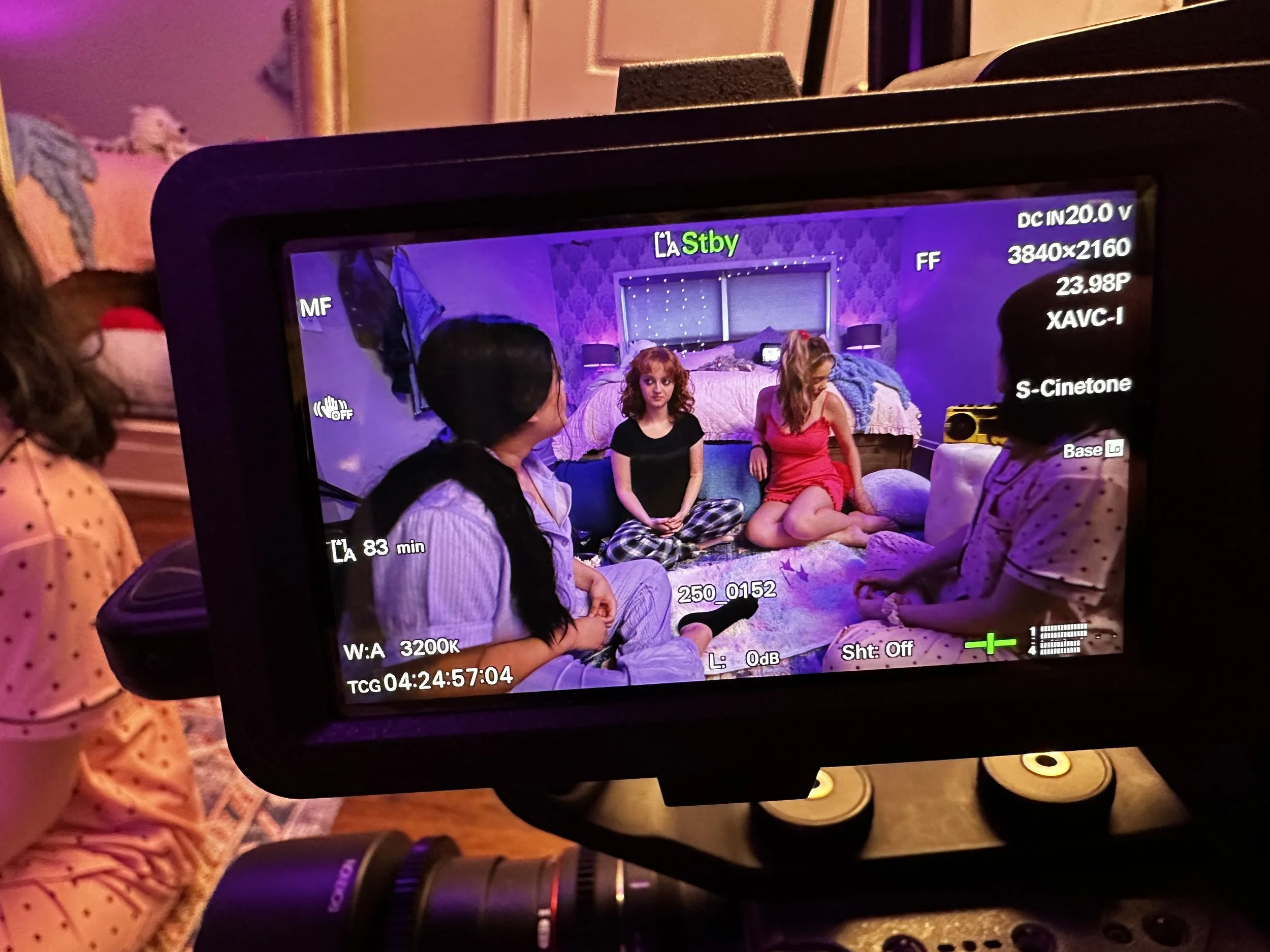 View of a camera screen displaying a scene with four people sitting on the floor in a cozy room with soft lighting and a bed in the background.