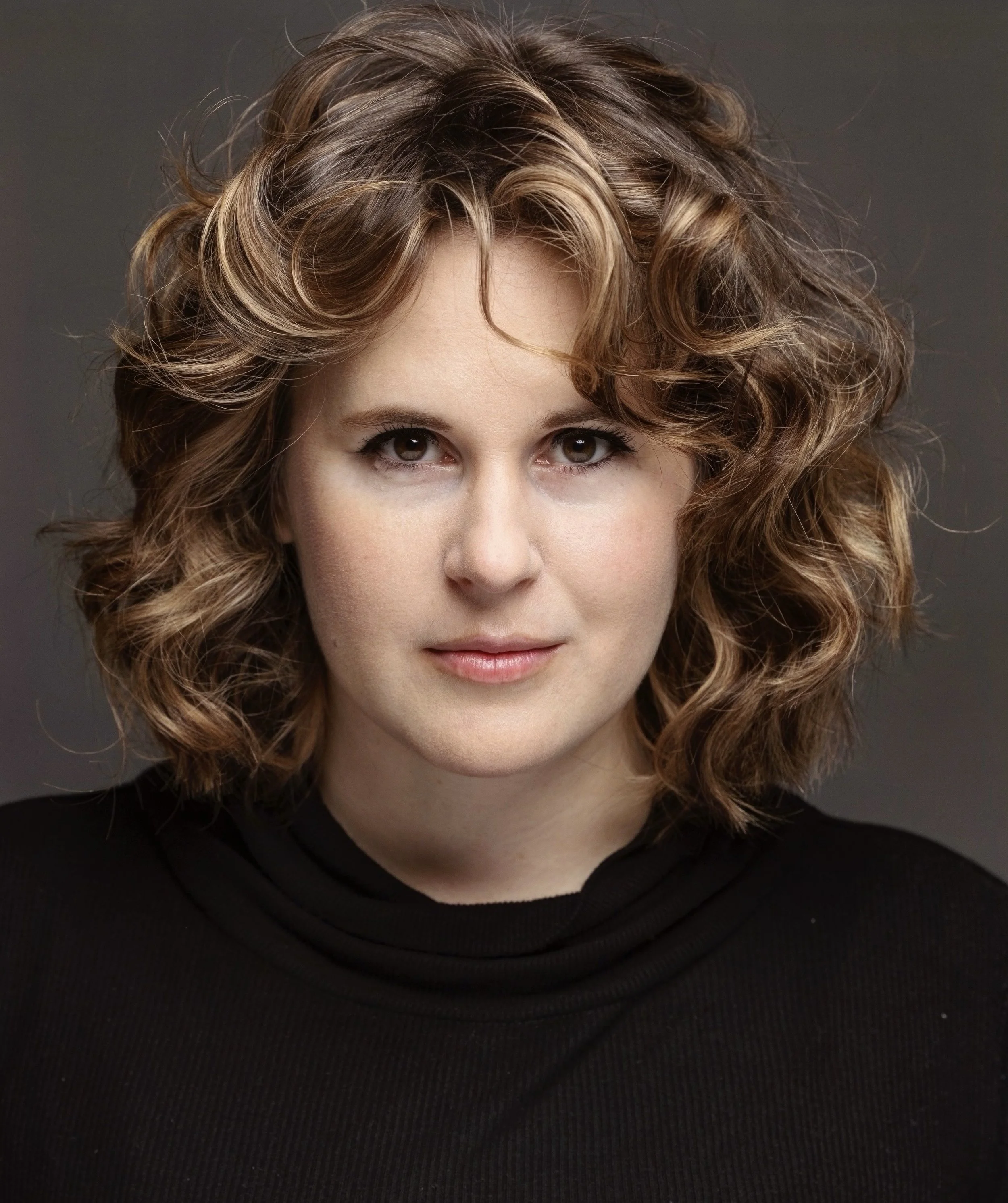 Headshot of actor with curly hair in black turtleneck against gray background.