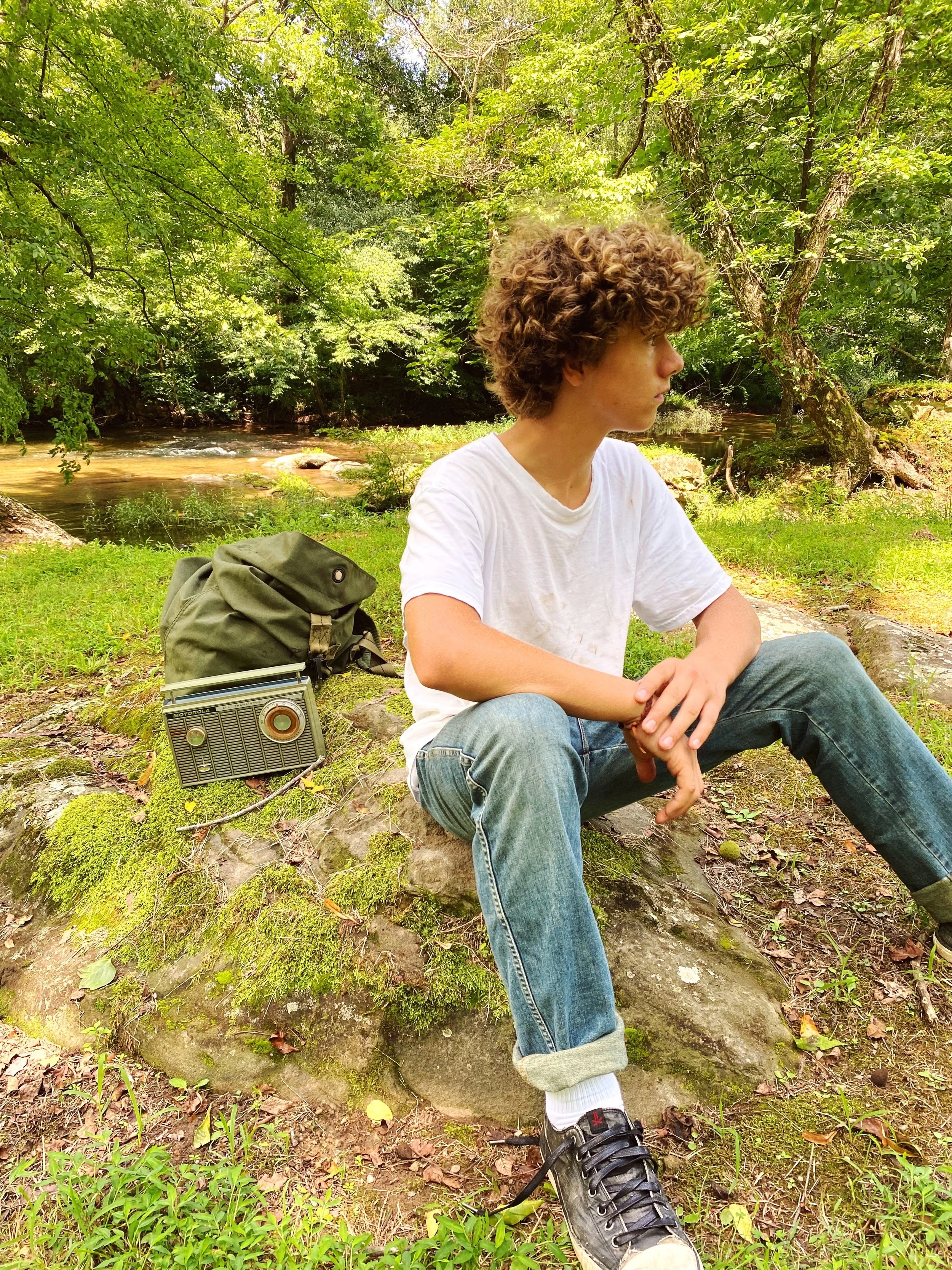 Person sitting on a moss-covered rock in a wooded area with a backpack and vintage radio nearby.
