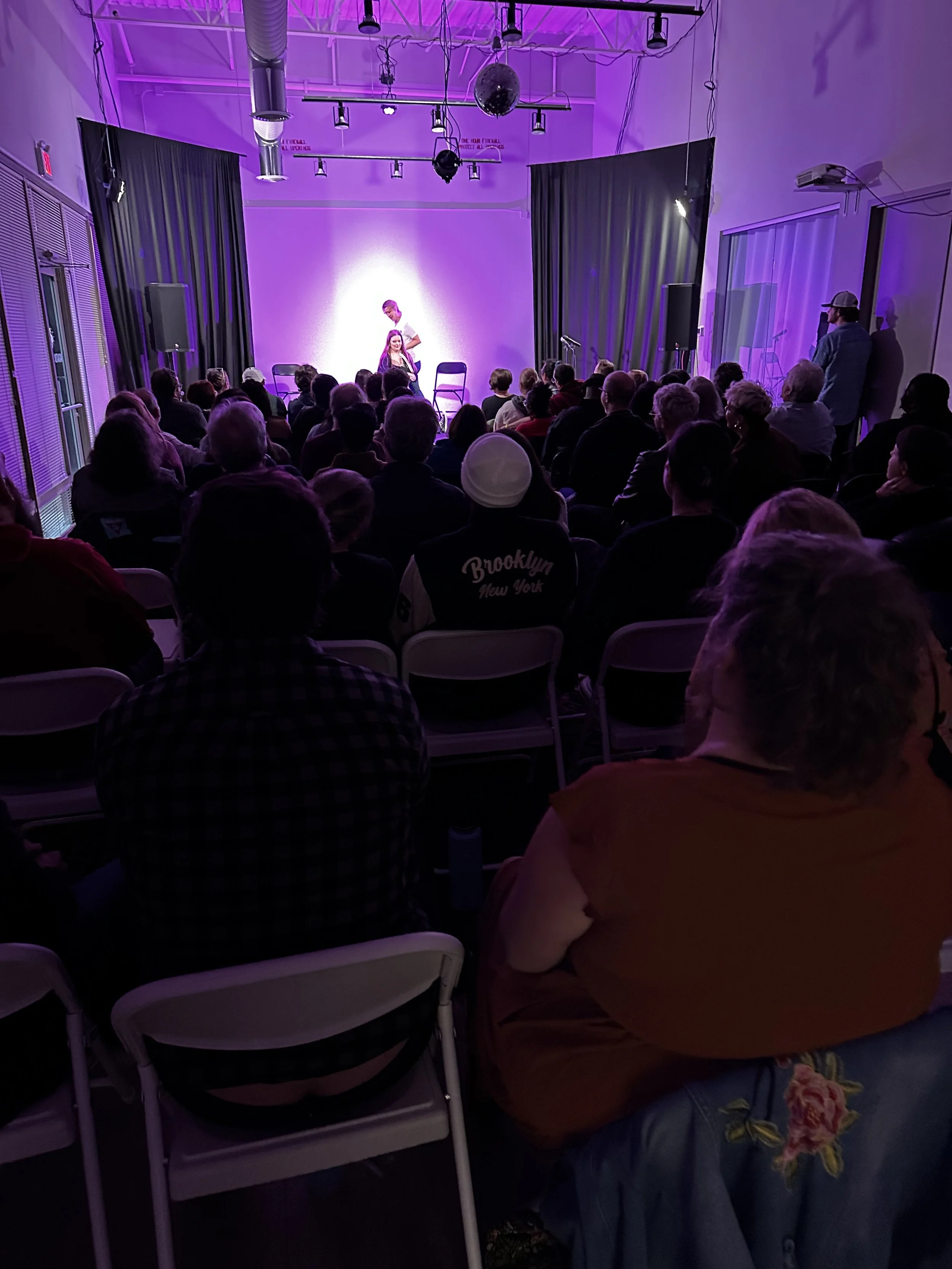 Audience watching a stage performance in a small venue with purple lighting.