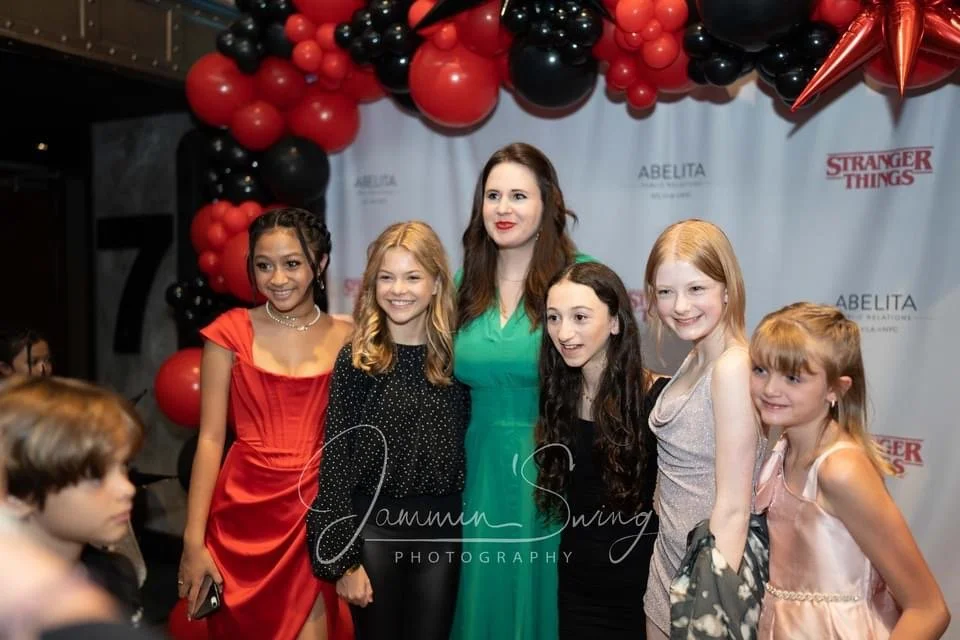 Group photo of six people at an event with a 'Stranger Things' backdrop, surrounded by red and black balloons. They are dressed in formal attire, smiling for the camera.