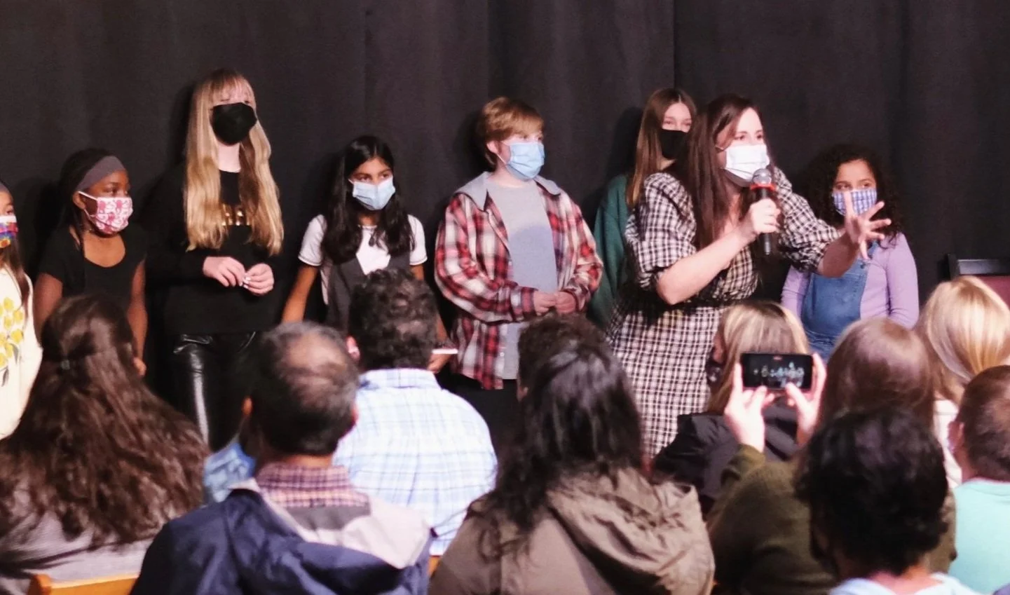 Group of children on stage with an adult speaking into a microphone, all wearing masks, facing an audience in a theater setting.