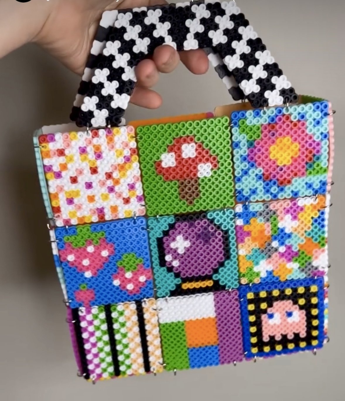 A colorful handbag made from small beads arranged in pixel art designs, featuring patterns like strawberries, a mushroom, a flower, and a retro game character.