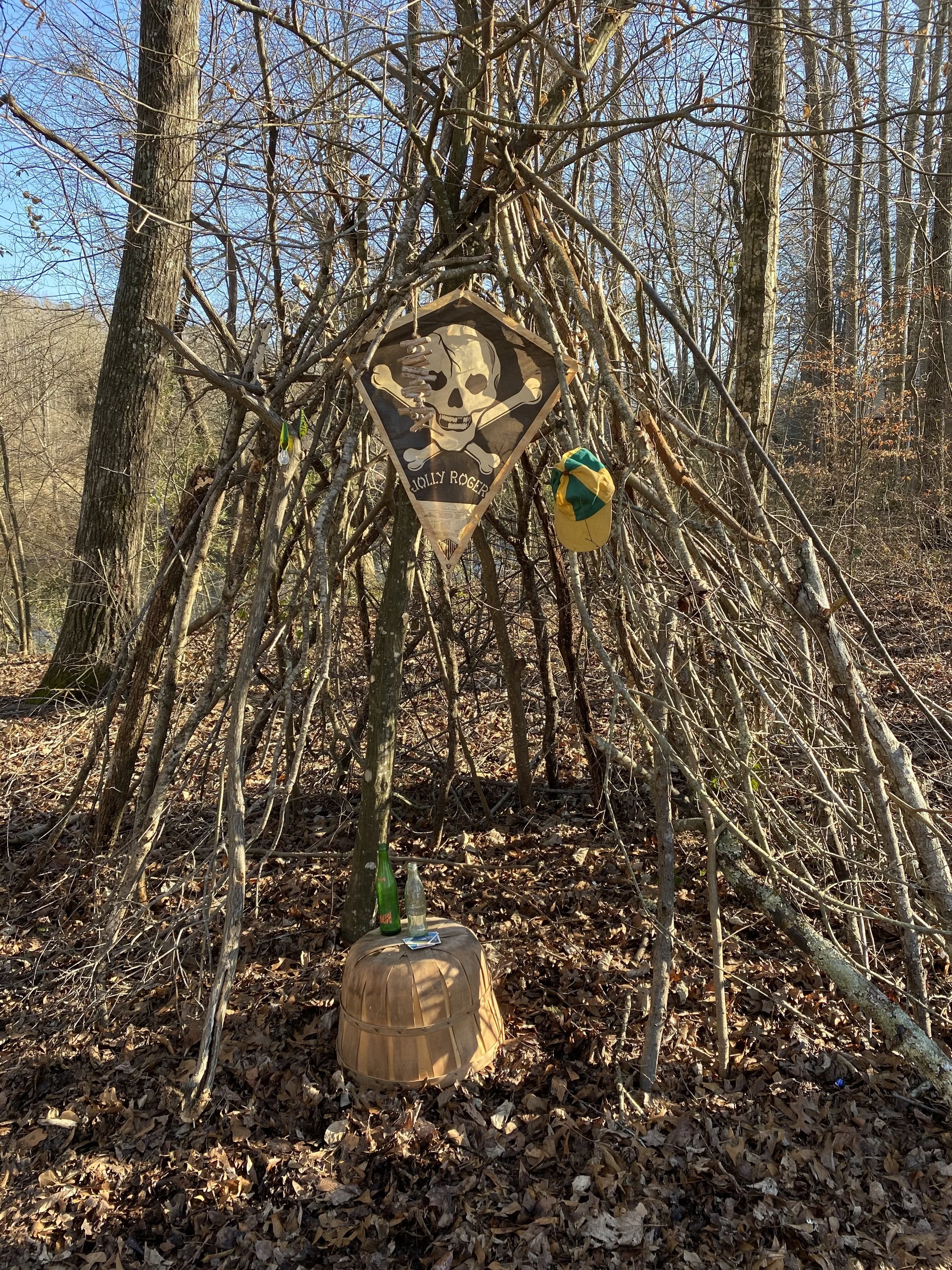 Teepee made of sticks in a forest with a Jolly Roger flag, yellow and green cap hanging, and small barrel with bottles on top on the ground.