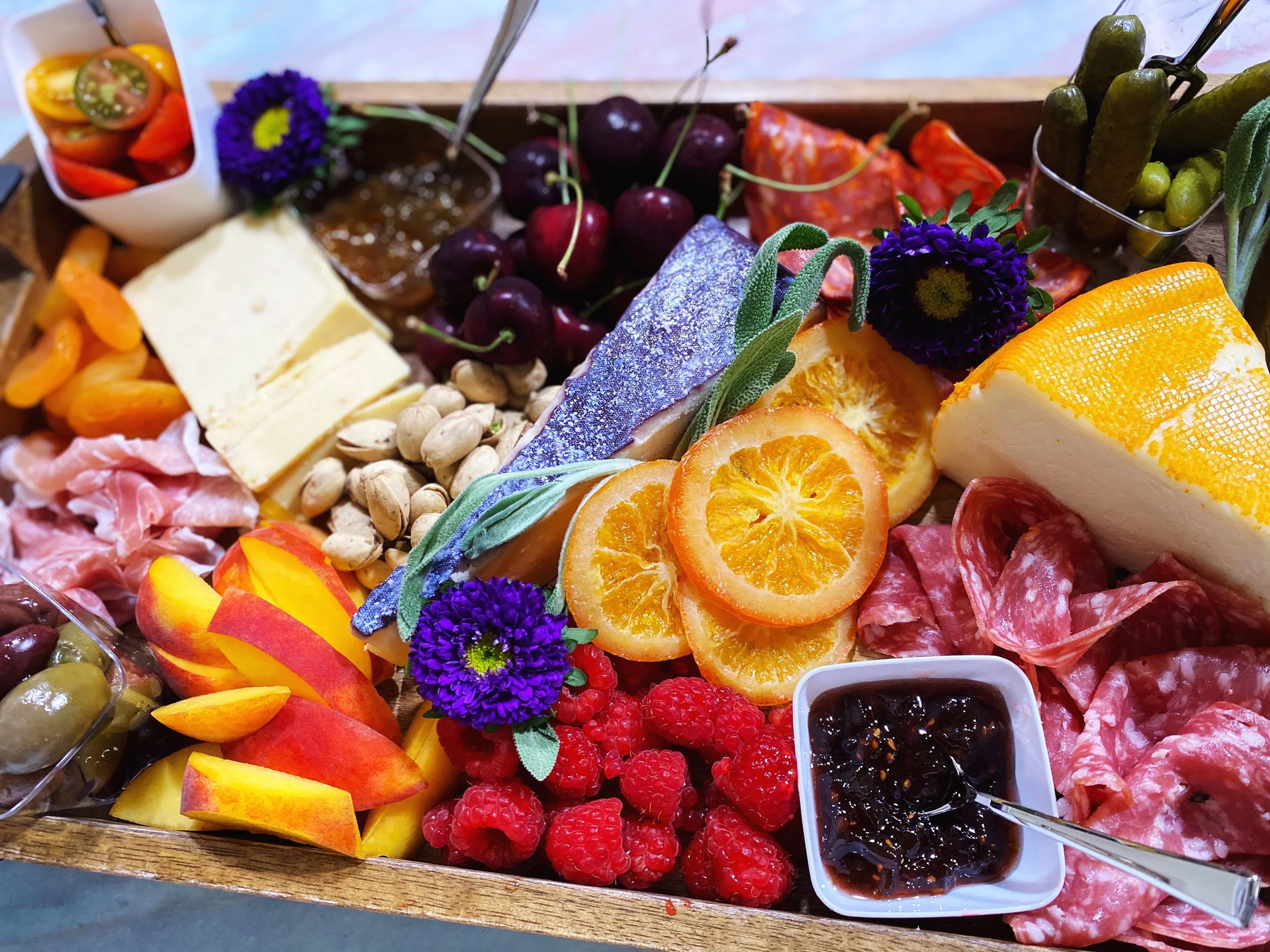 Charcuterie board with assorted cheeses, sliced salami, prosciutto, dried apricots, pistachios, fresh cherries, blackberries, raspberries, sliced peaches, pickles, orange slices, purple flowers, and dipping sauces in small containers, garnished with green herbs.