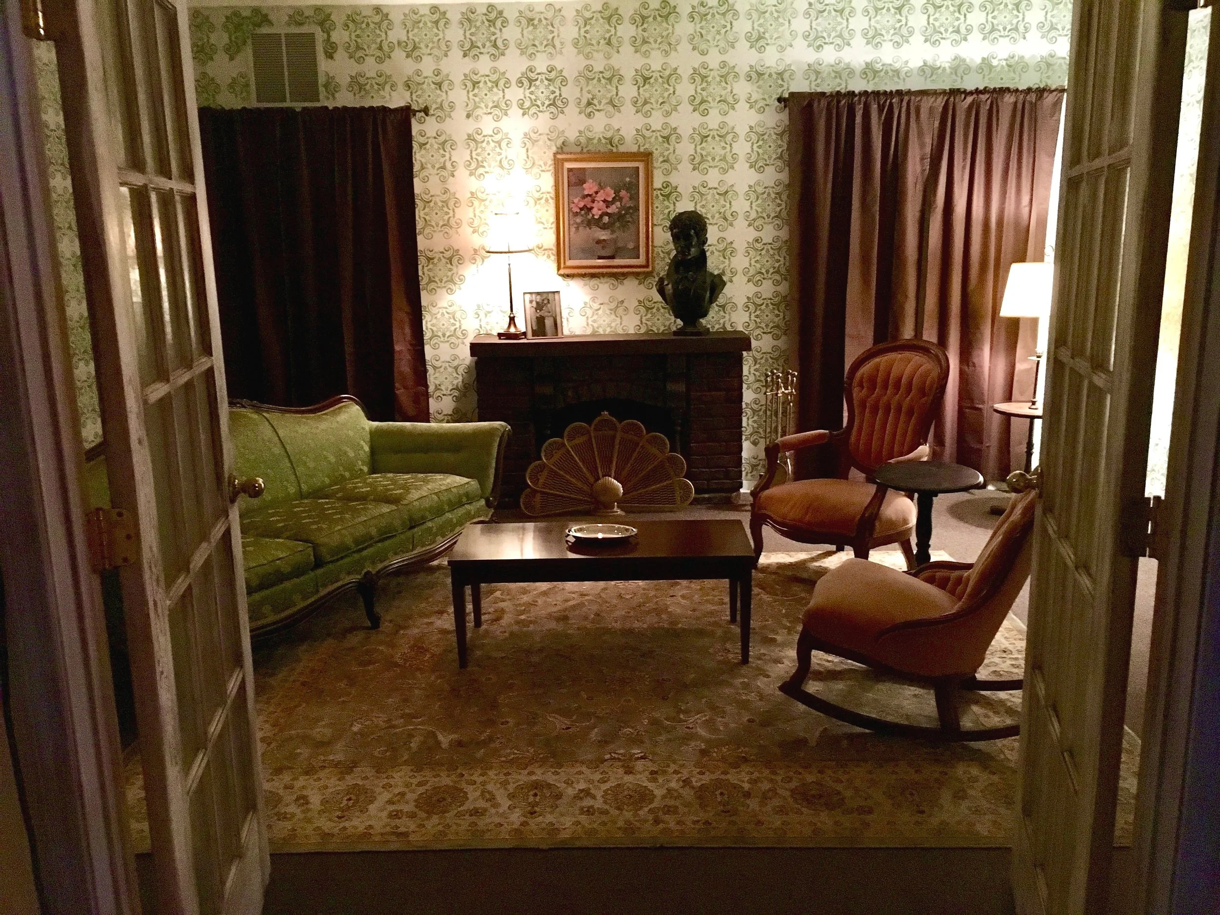 Vintage living room with green couch, decorative armchairs, dark drapes, patterned wallpaper, and a fireplace with artwork and a lamp.