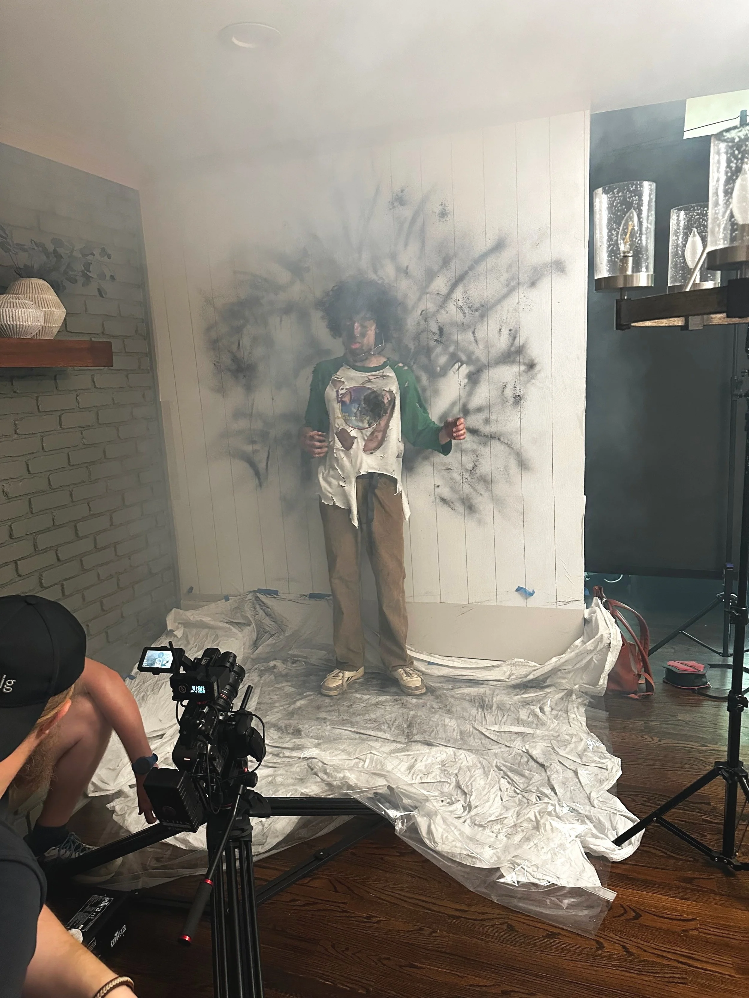 A person stands against a white panel wall covered in soot or black marks, surrounded by hazy smoke. They wear a green-sleeved shirt and khaki pants. In front of them is a professional camera setup with a person operating it. The floor is covered with protective sheets. The scene suggests a video or photo shoot, possibly involving special effects.