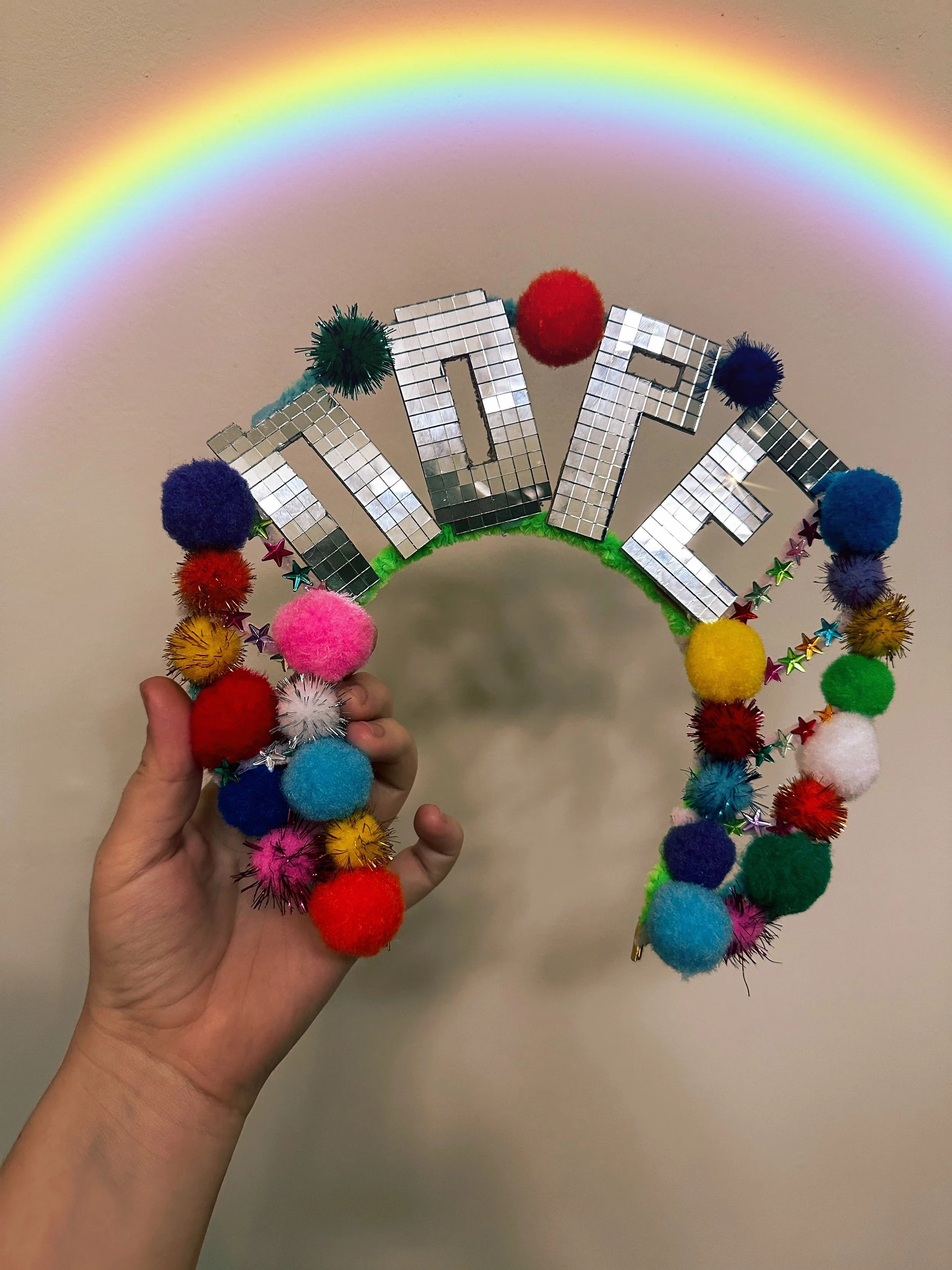 A hand holding a decorative two layered angel halo headband covered with colorful pom-poms, mirrored letters spelling "NOPE" and a rainbow overhead.