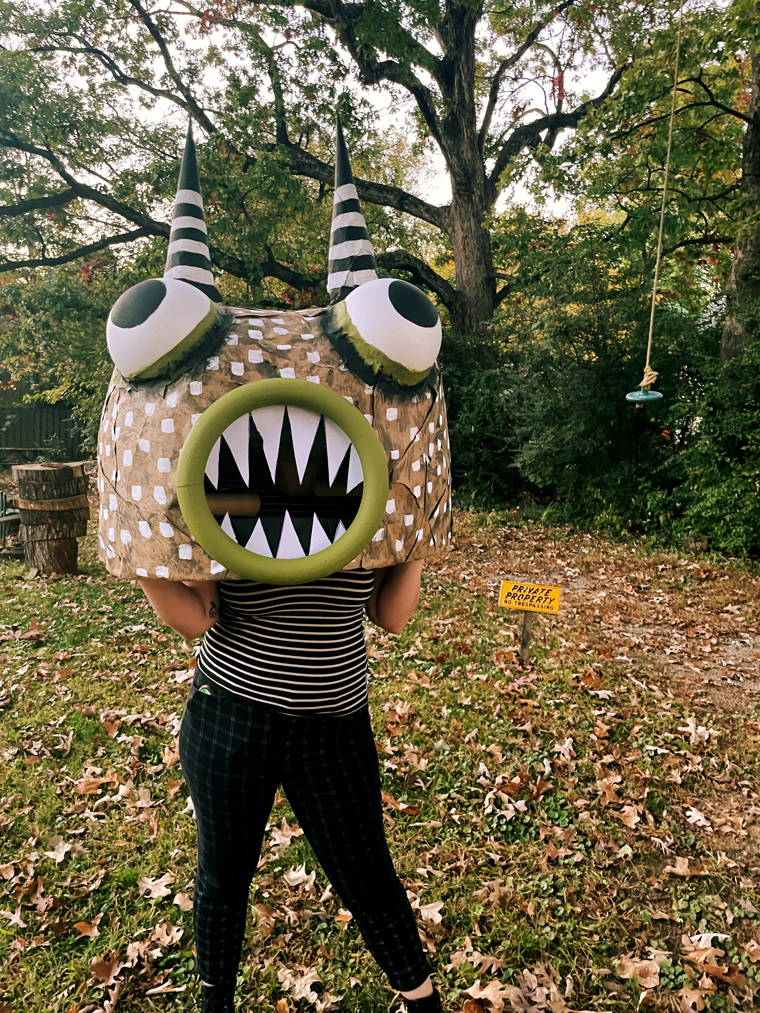 Person wearing large monster head costume in outdoor setting with trees and autumn leaves.