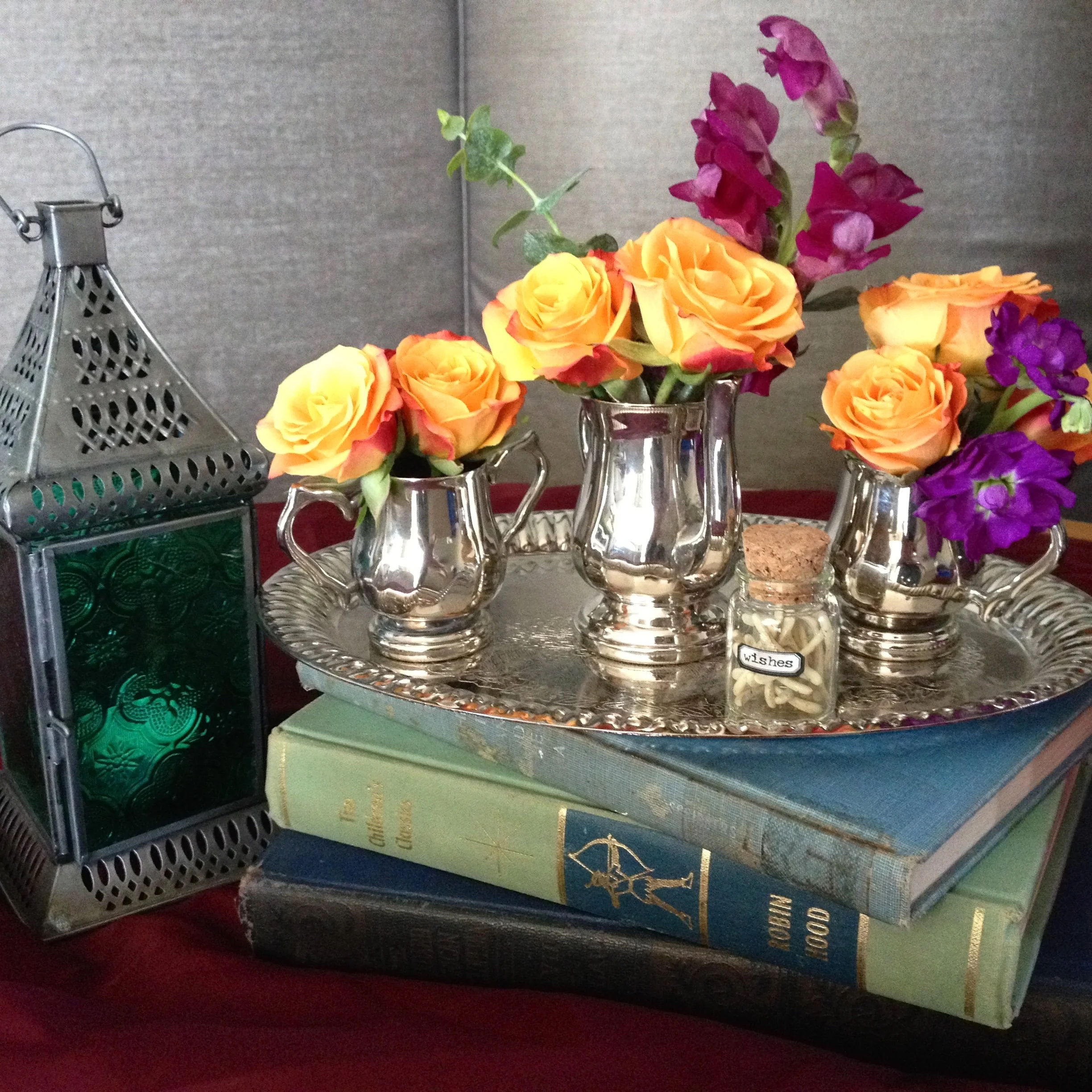 A decorative arrangement featuring a silver tray with small silver vases holding orange roses and purple flowers, alongside a small glass jar labeled "wishes." The tray is placed on a stack of vintage books and next to an ornate metal lantern with gr