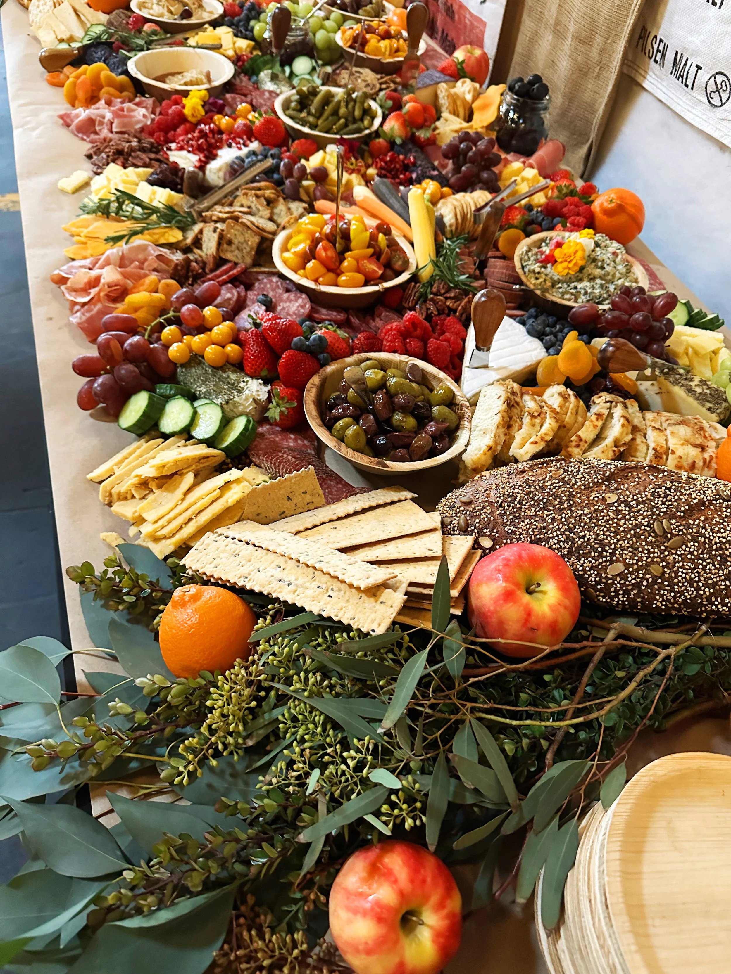 A colorful charcuterie board with a variety of cheeses, meats, fruits, vegetables, bread, olives, and crackers, arranged with decorative greenery and apples.