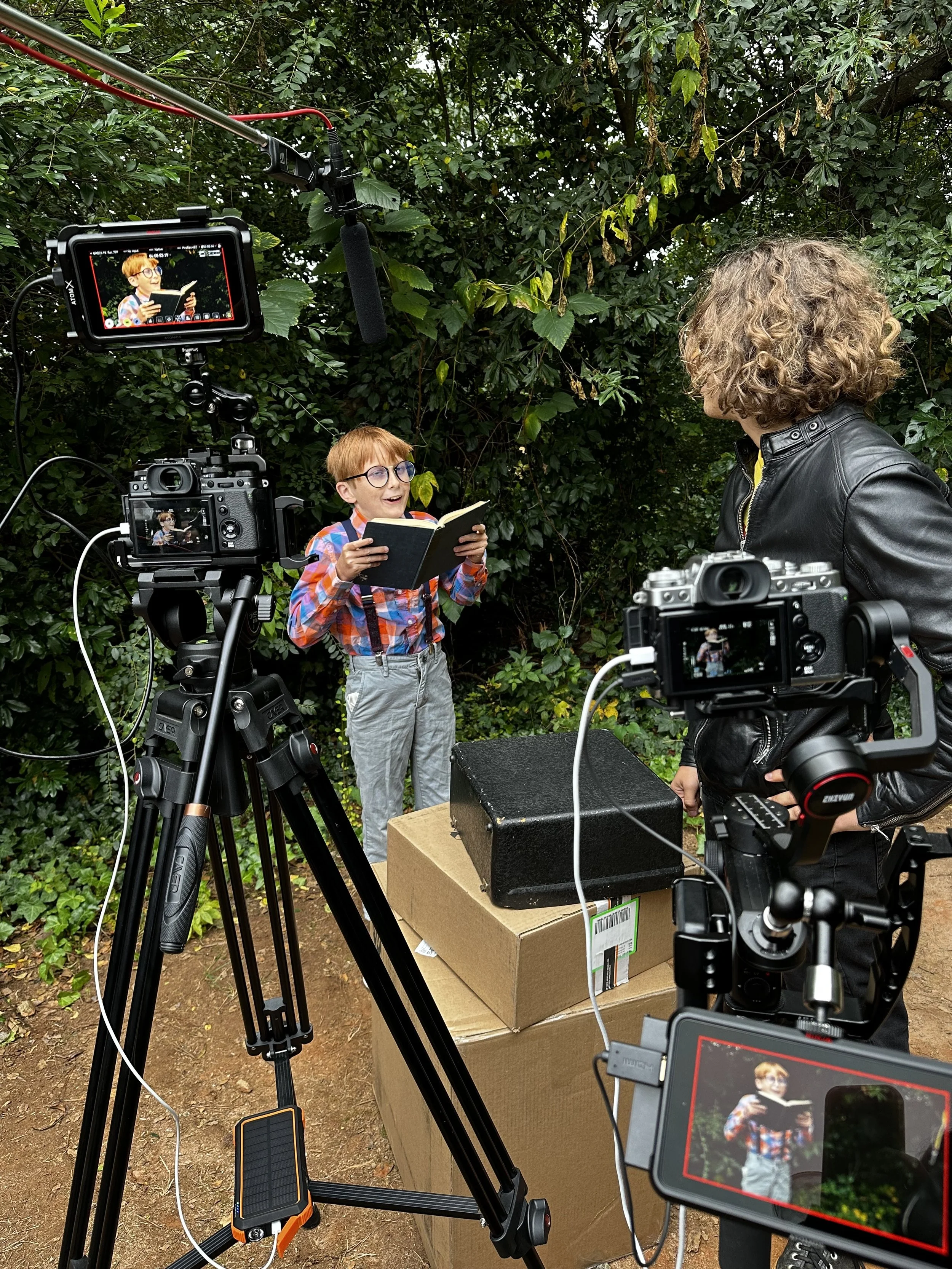 A film set scene in a forest, featuring a person wearing a plaid shirt and glasses, reading from a book. They're surrounded by filming equipment, including cameras on tripods, microphones, and monitors displaying the scene. Another person with curly hair is visible, engaging with the filming process.