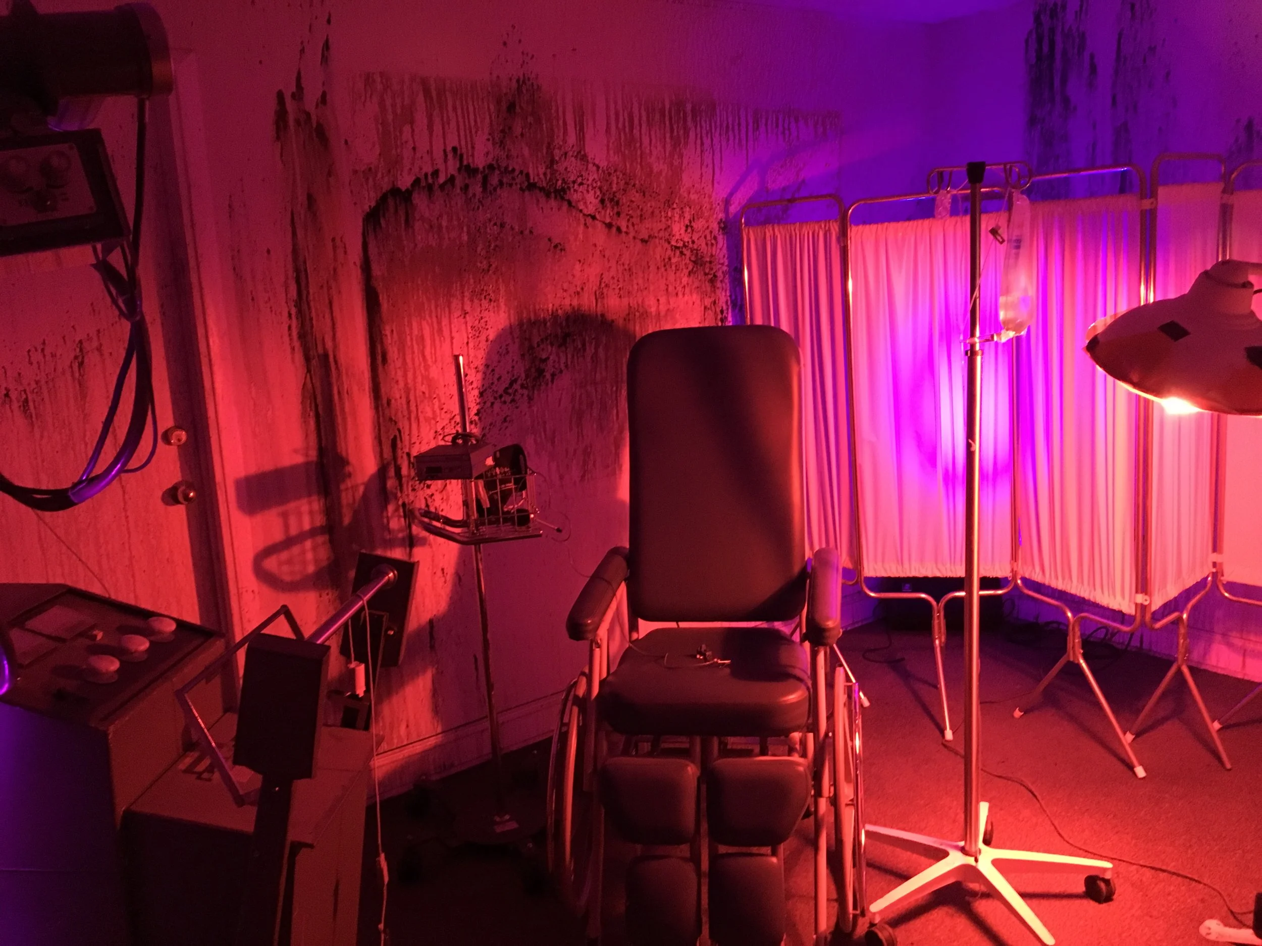 A dimly lit room with a medical chair, some medical equipment, and a privacy screen, illuminated by red and purple lights.