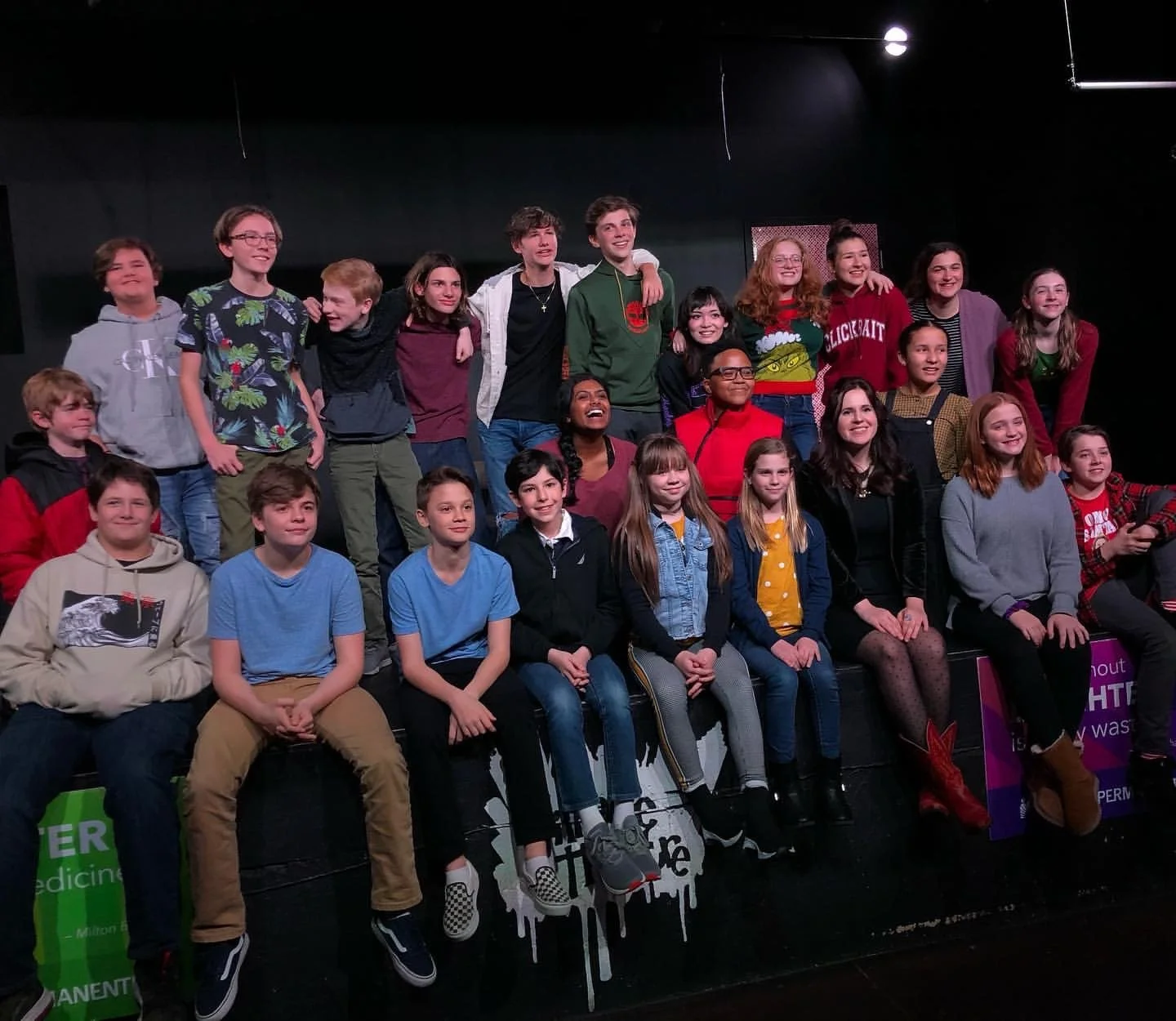 Group of teenagers and a few adults posing together, some seated and others standing, on a stage with a dark background. They are happy after a theatrical event.