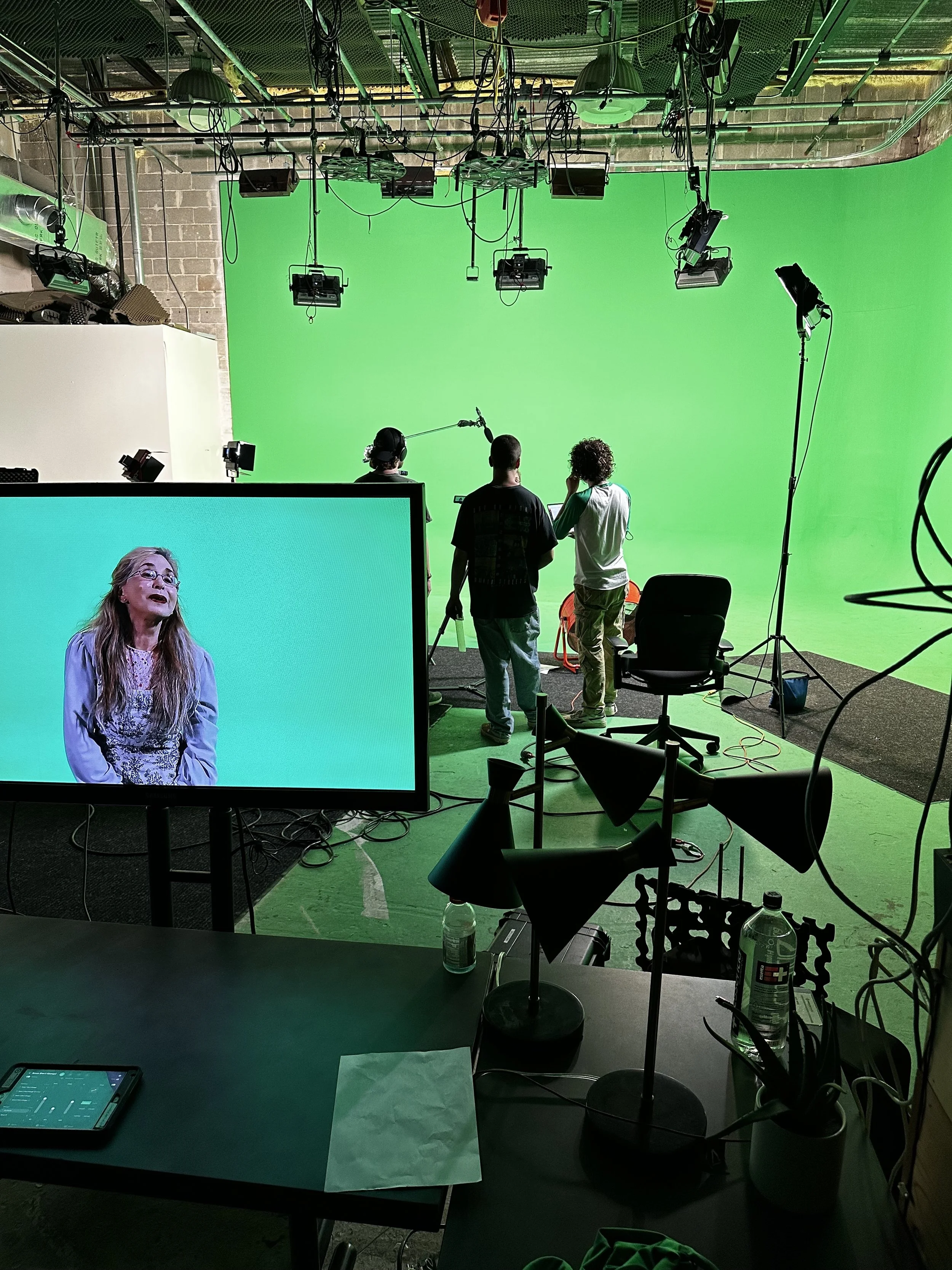 A green screen studio setup with lighting and equipment. Three people are standing in front of the green screen. A monitor shows a person in front of a green background. There are lamps and a water bottle on a table in the foreground.