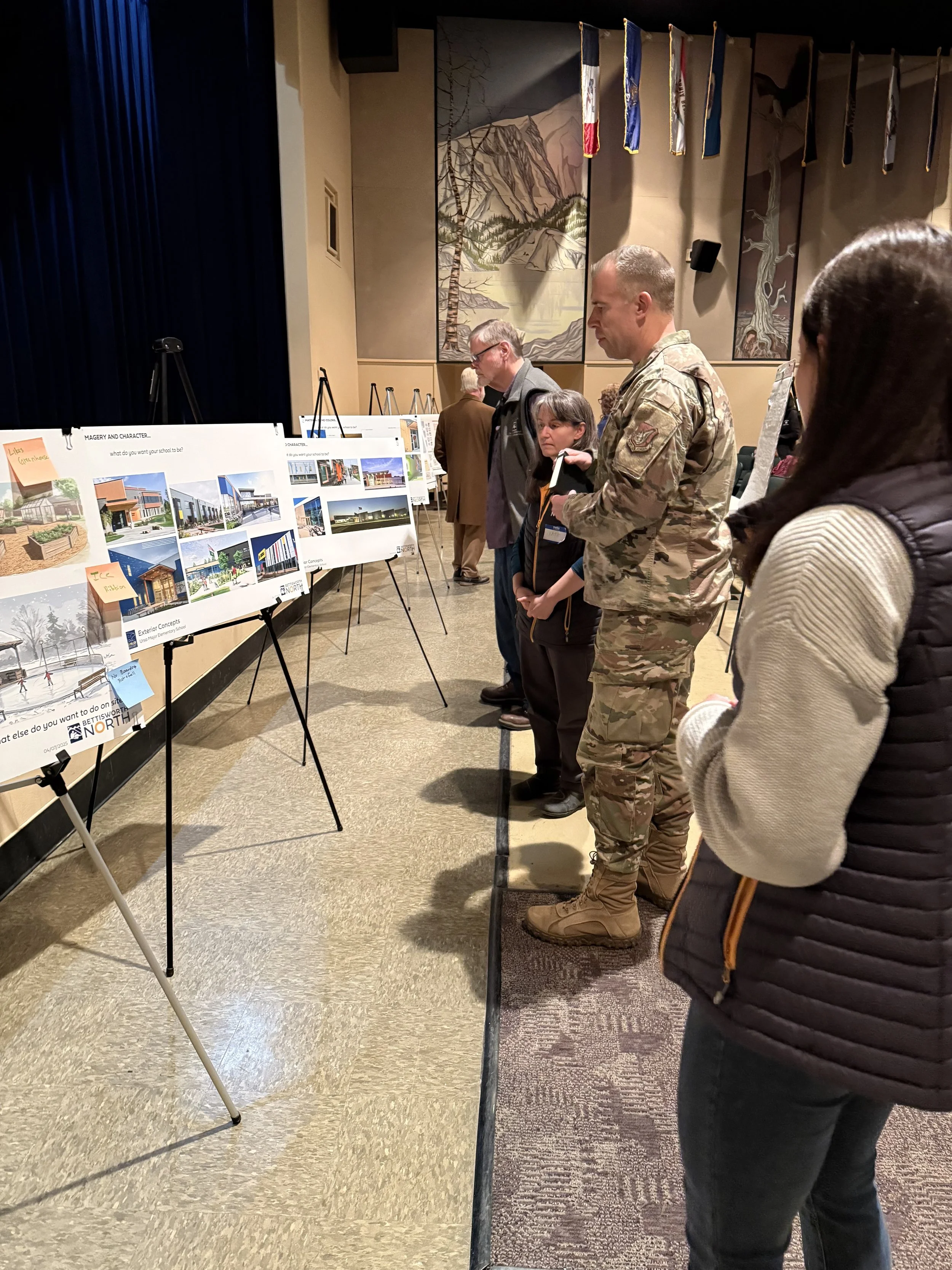 People in a room viewing architectural design boards displayed on easels, with one person in military uniform. The room features a mural and flags hanging on the walls.