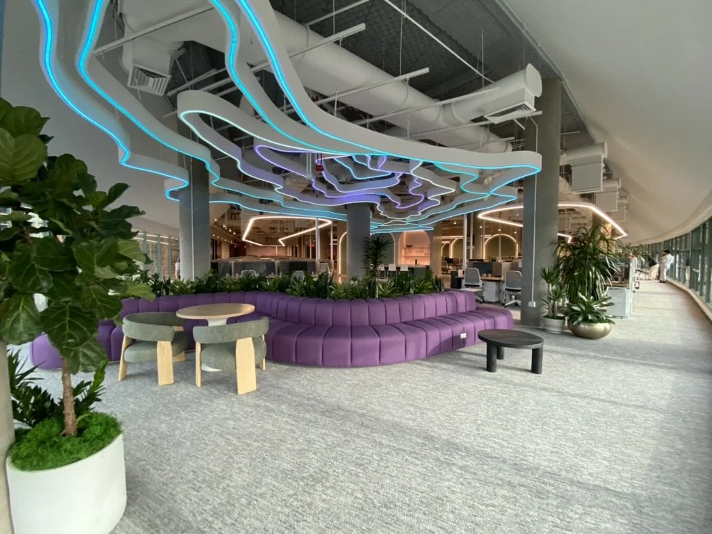 A modern office lounge with a curved purple sofa, lush indoor plants, and wave-like LED ceiling lights, set beside floor-to-ceiling windows.