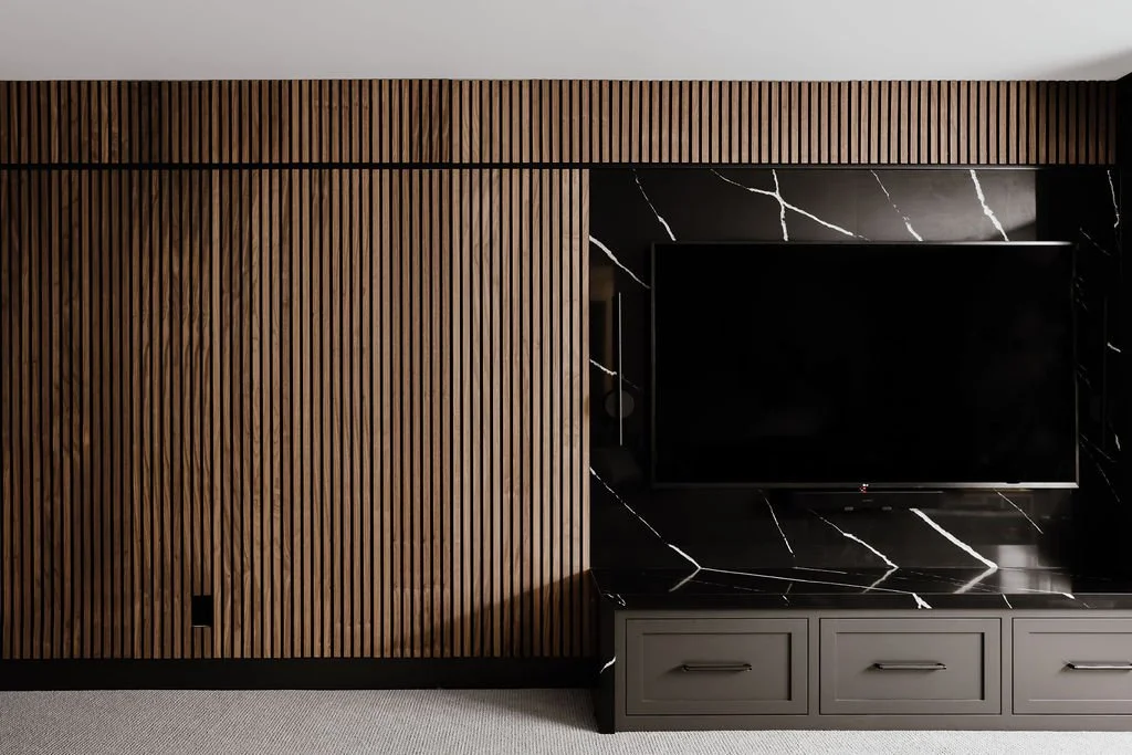 Walnut slat wall, wall mounted tv with black quartz behind it, dark gray bench cabinets below the tv, bench made from the same black quartz with a waterfall edge. Sconce next to the tv. 