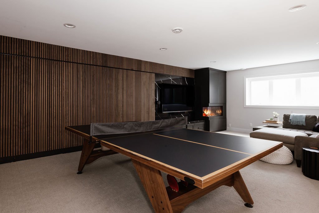 Wide shot of a finished basement with a window, walnut slat wall, electric holographic fireplace, wall mounted tv with black quartz behind it, ping pong table and furniture seating. 