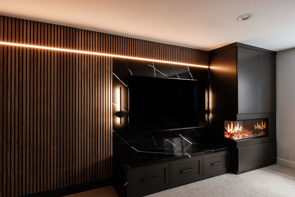 Walnut slat wall now showing the black horizontal line is a LED strip light, electric holographic fireplace, wall mounted tv with black quartz behind it, two lit up wall sconces flanking the tv. 