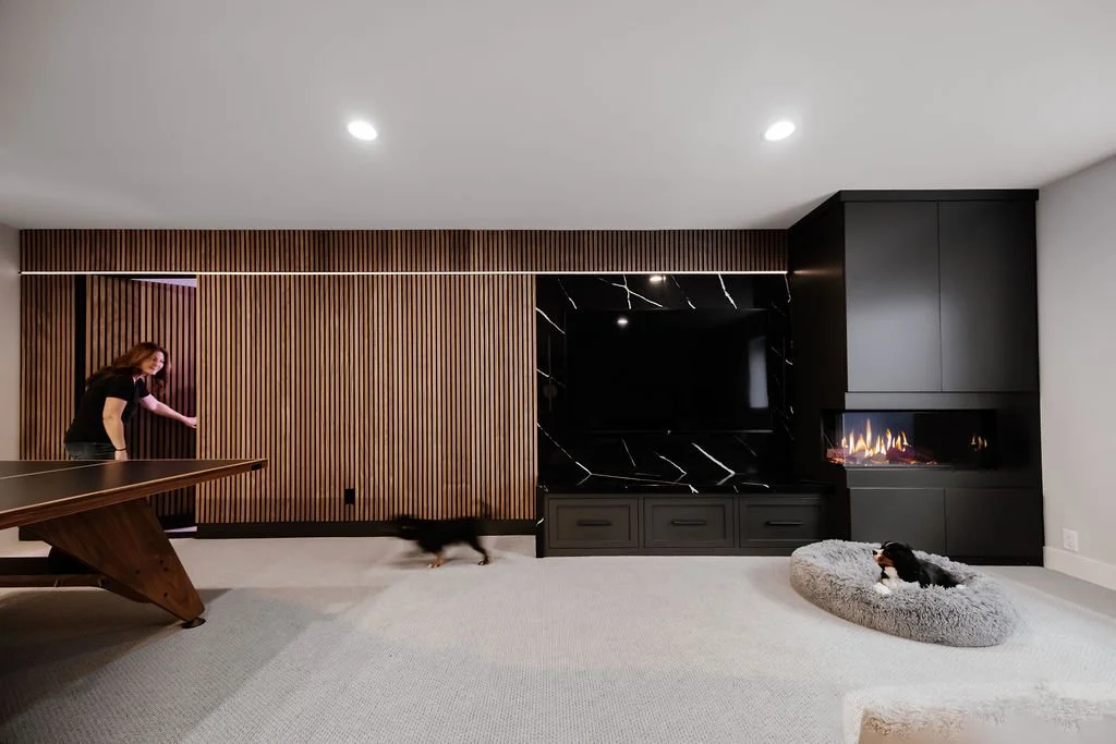 Wide shot of the basement area showing the accent wall with the owner and general contractor Rachel Schaefer and her two King Charles Cavalier Spaniel Dogs, Otis and Bernie. Rachel has opened the walnut slat wall's hidden door into a storage room. Ot