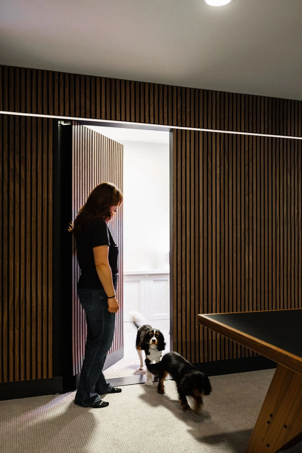 The slat wall door is open and you can see a peek of a storage room with white base cabinetry. Otis and Bernie are coming out of the storage area into the main basement room while Rachel holds the door open. 