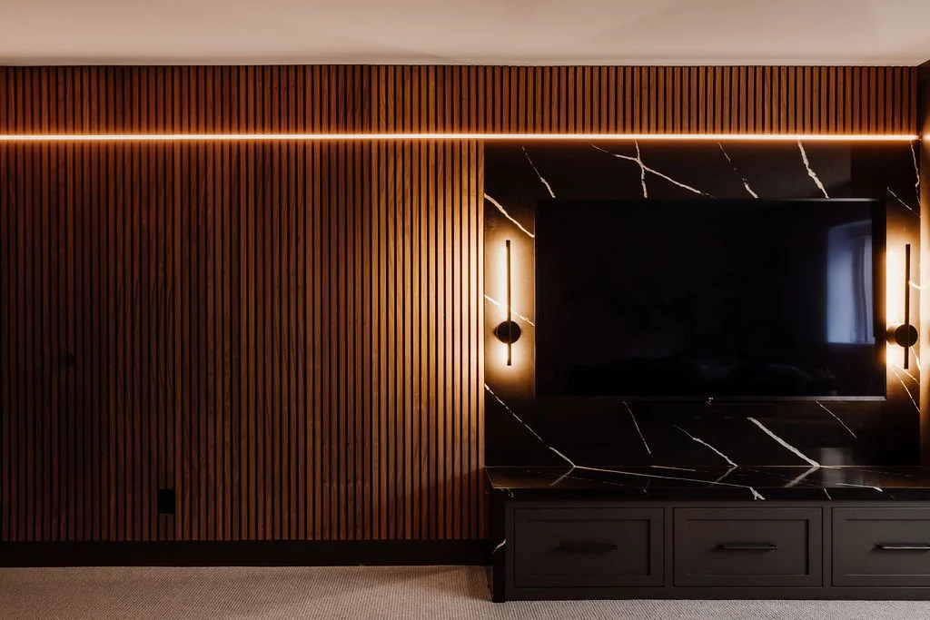 A front angle of the tv area with walnut slat wall, LED strip light, wall mounted tv with black quartz behind it, two lit up wall sconces flanking the tv. 