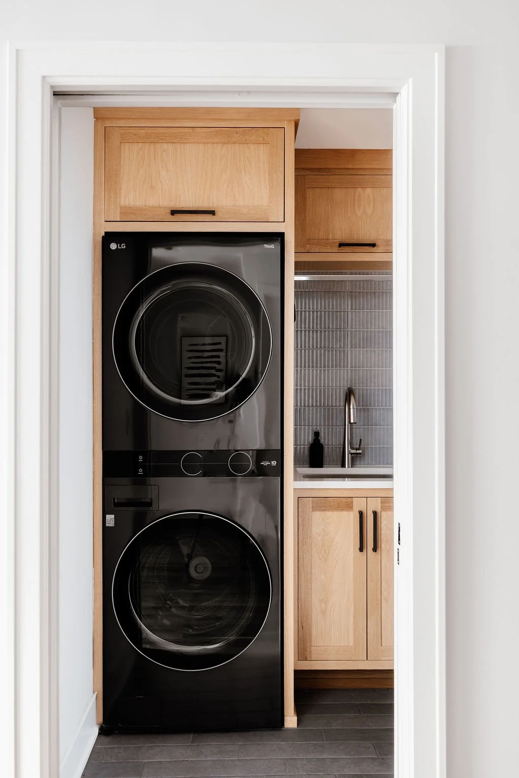 Small modern laundry room with graphite color wash/dry tower, Rift white oak cabinetry to the ceiling including a spot above the wash/dry tower for laundry hamper. Deep utility sink, black and stainless accents, clothes hanging rod above the sink and