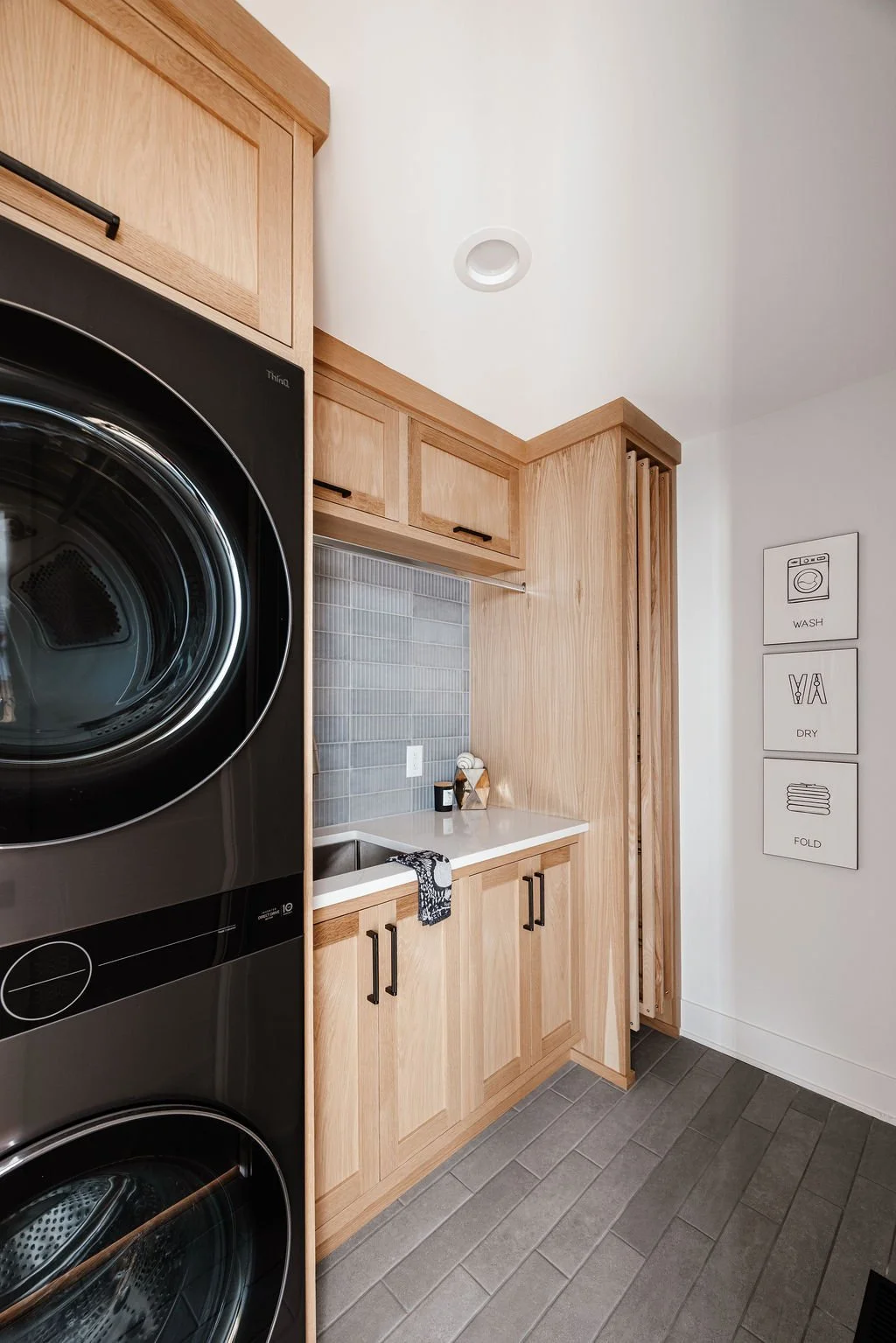 Small modern laundry room with graphite color wash/dry tower, Rift white oak cabinetry to the ceiling including a spot above the wash/dry tower for laundry hamper. Deep utility sink, black and stainless accents, clothes hanging rod above the sink and