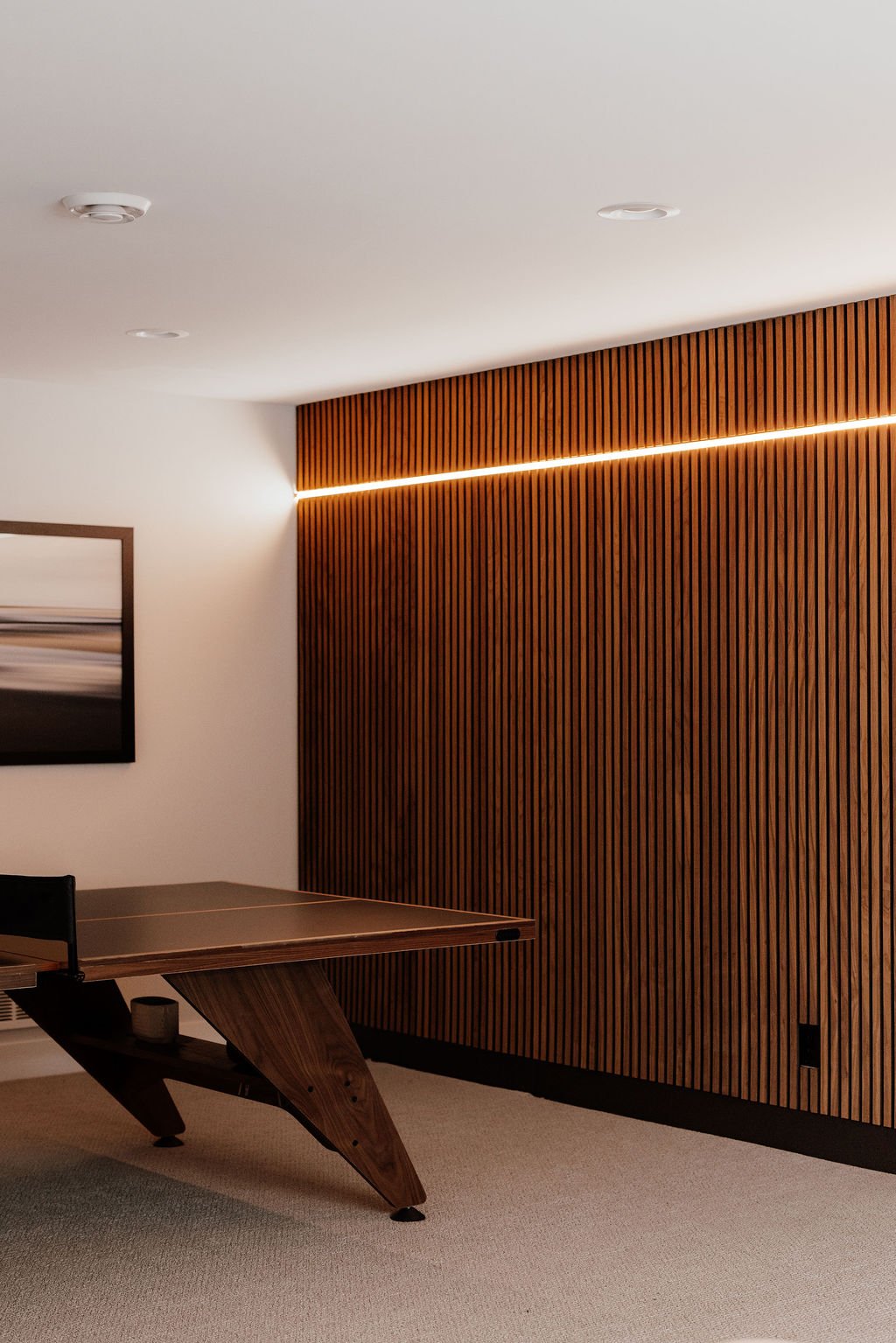 Modern basement feature wall with walnut acoustic slat panels, integrated LED lighting, framed art, and a walnut ping-pong table creating a warm, functional recreation space.