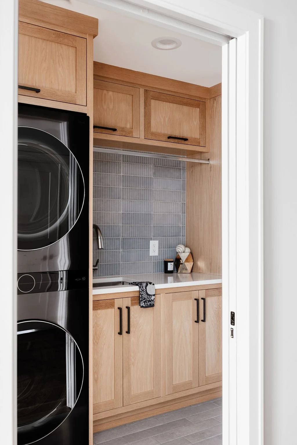 Small modern laundry room with graphite color wash/dry tower, Rift white oak cabinetry to the ceiling including a spot above the wash/dry tower for laundry hamper. Deep utility sink, black and stainless accents, clothes hanging rod above the sink and