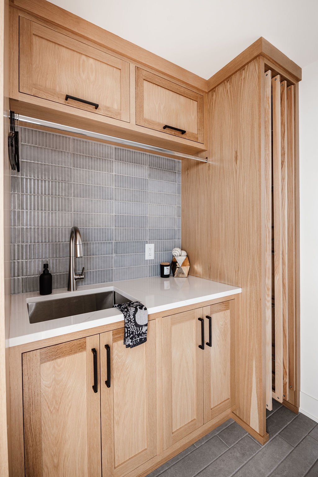 Small modern laundry room with rift white oak cabinetry to the ceiling, deep stainless utility sink, Delta Monrovia pull down stainless facuet, integrated drying racks, black cabinet pulls, light blue backsplash tile with texture.