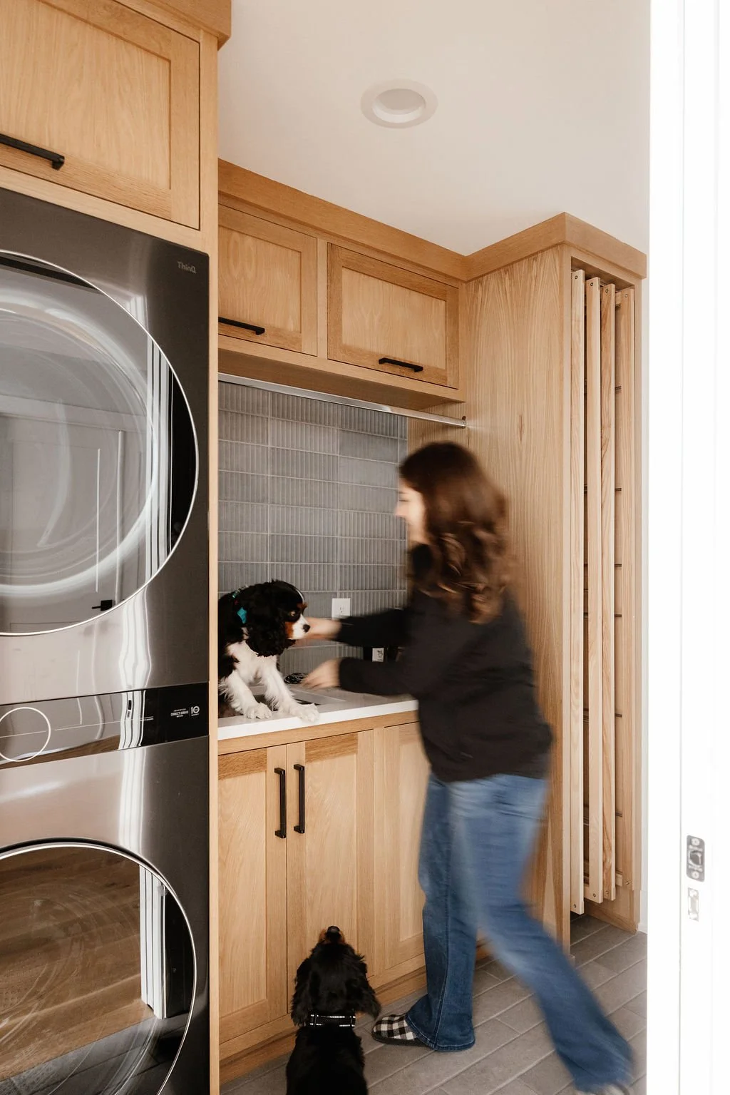 Small modern laundry room with stacked washer and dryer, Rift white oak cabinetry to the ceiling, deep utility sink, white quartz countertop, integrated drying racks, black and stainless accents, and a large pocket door. Rachel Schaefer puts her dog 