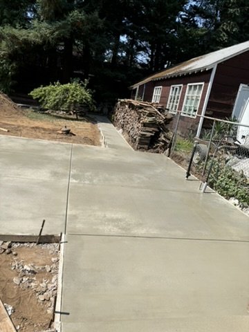 Freshly poured concrete walkway leading to a house with a woodpile, surrounded by trees.