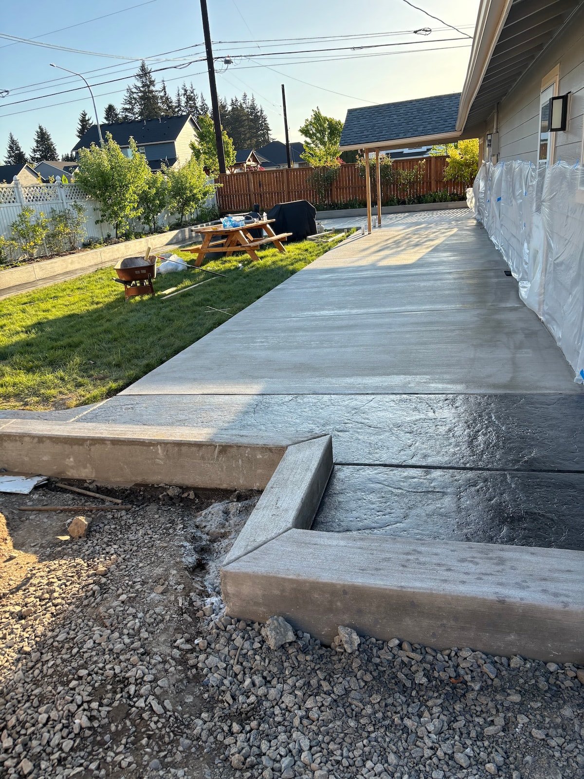 Backyard with a newly poured concrete patio, a picnic table on the grass, and a covered area near the house. The yard has a wooden fence and trees in the background.