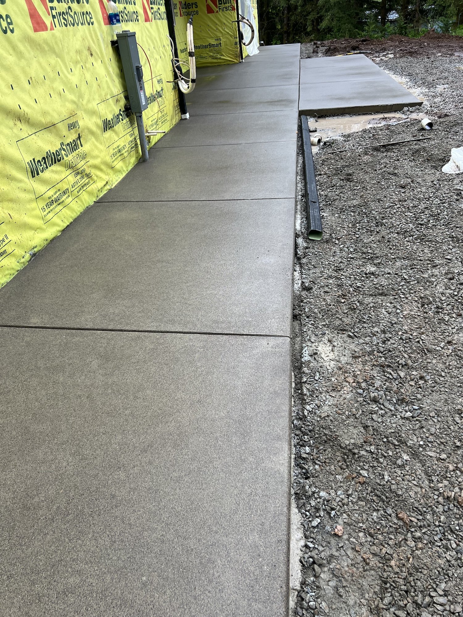 Newly poured concrete walkway beside a building with yellow WeatherSmart wrap, surrounded by gravel and dirt.