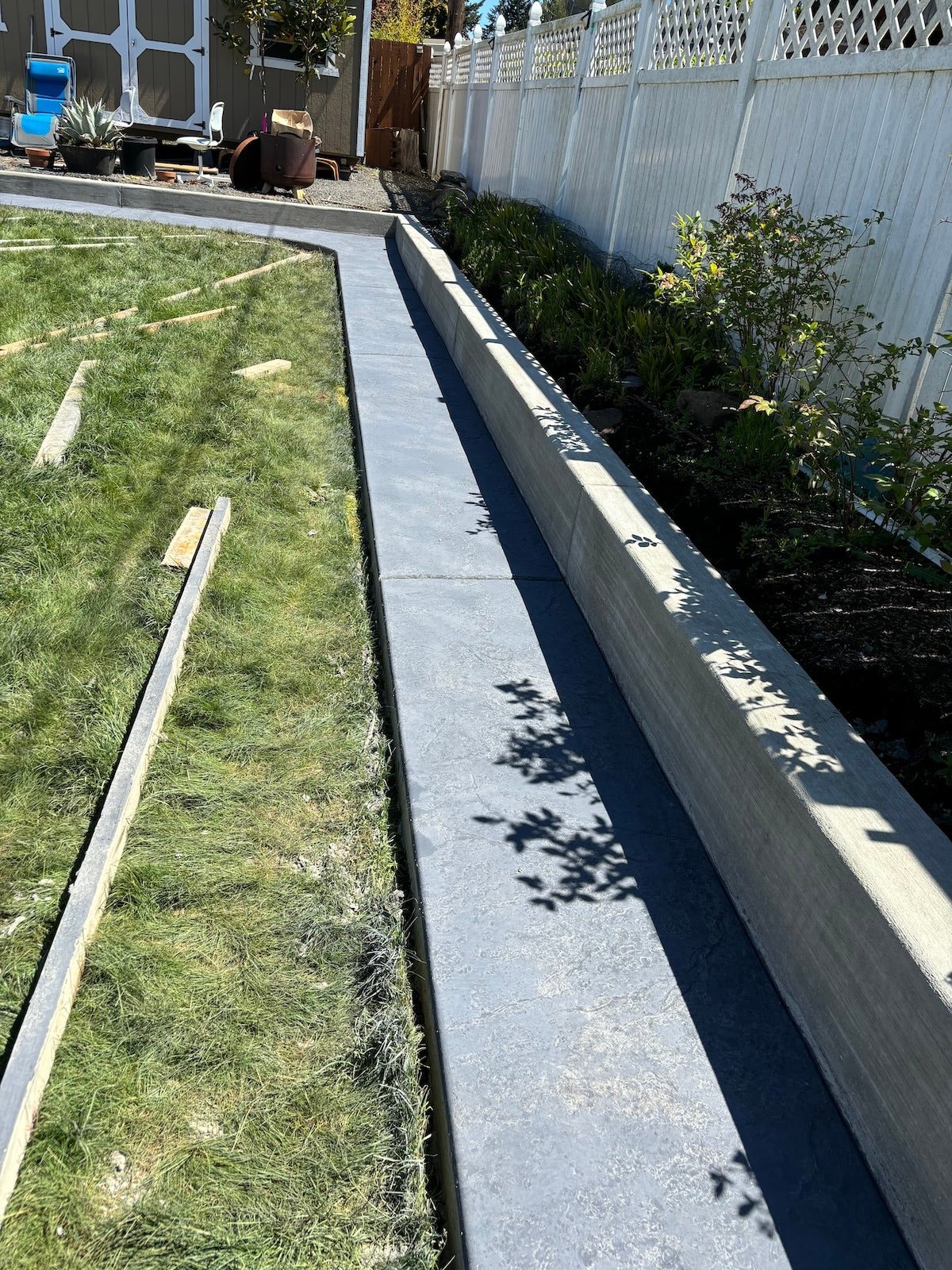 Backyard with newly constructed concrete path, adjacent grass and garden bed, bordered by a white fence. Patio furniture in the background.